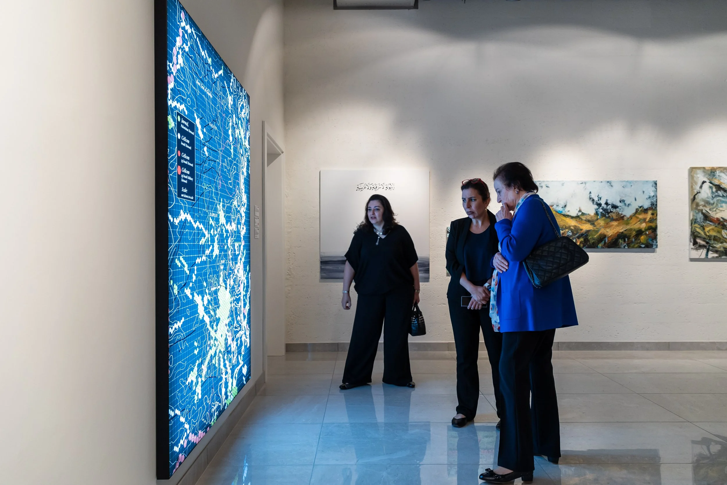 Three women are standing in an art gallery and viewing a digital display on a wall, with paintings hanging behind them.