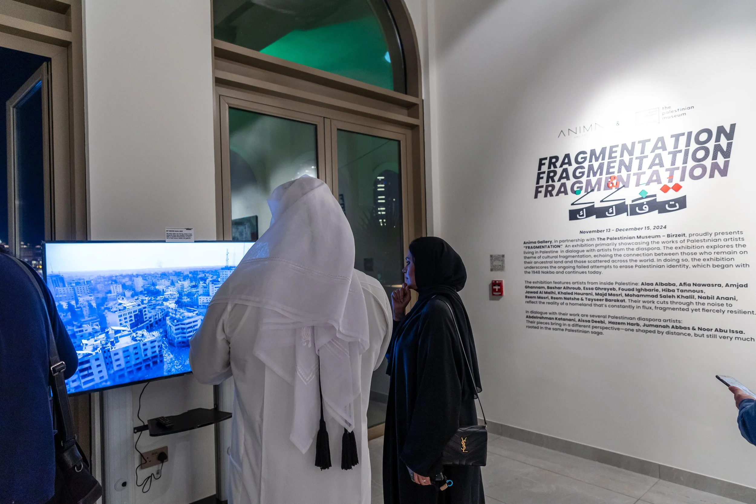 Two women in headscarves looking at a large digital screen displaying an aerial view of a city, in a gallery with text about an exhibition called 'Fragmentation' at the Palestinian Museum.