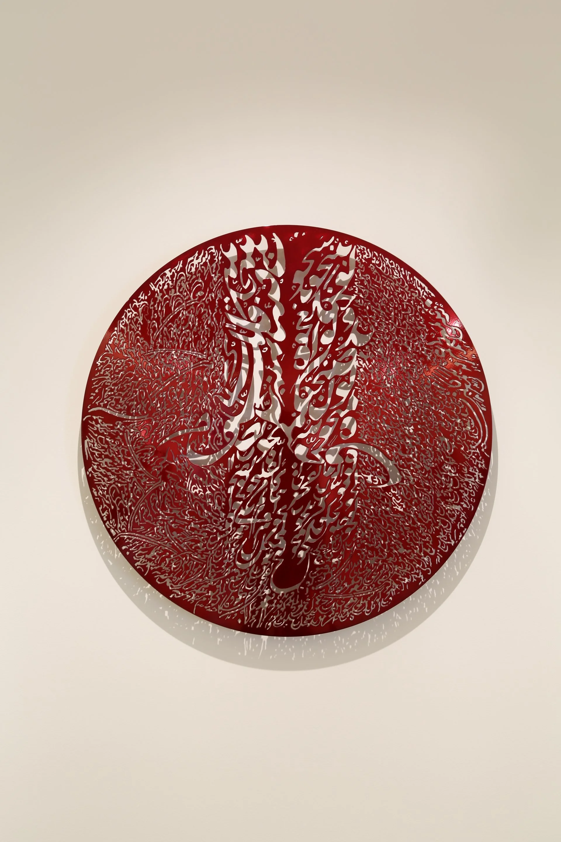 Intricate circular red artwork with Arabic calligraphy and designs, casting a shadow on a plain white wall.