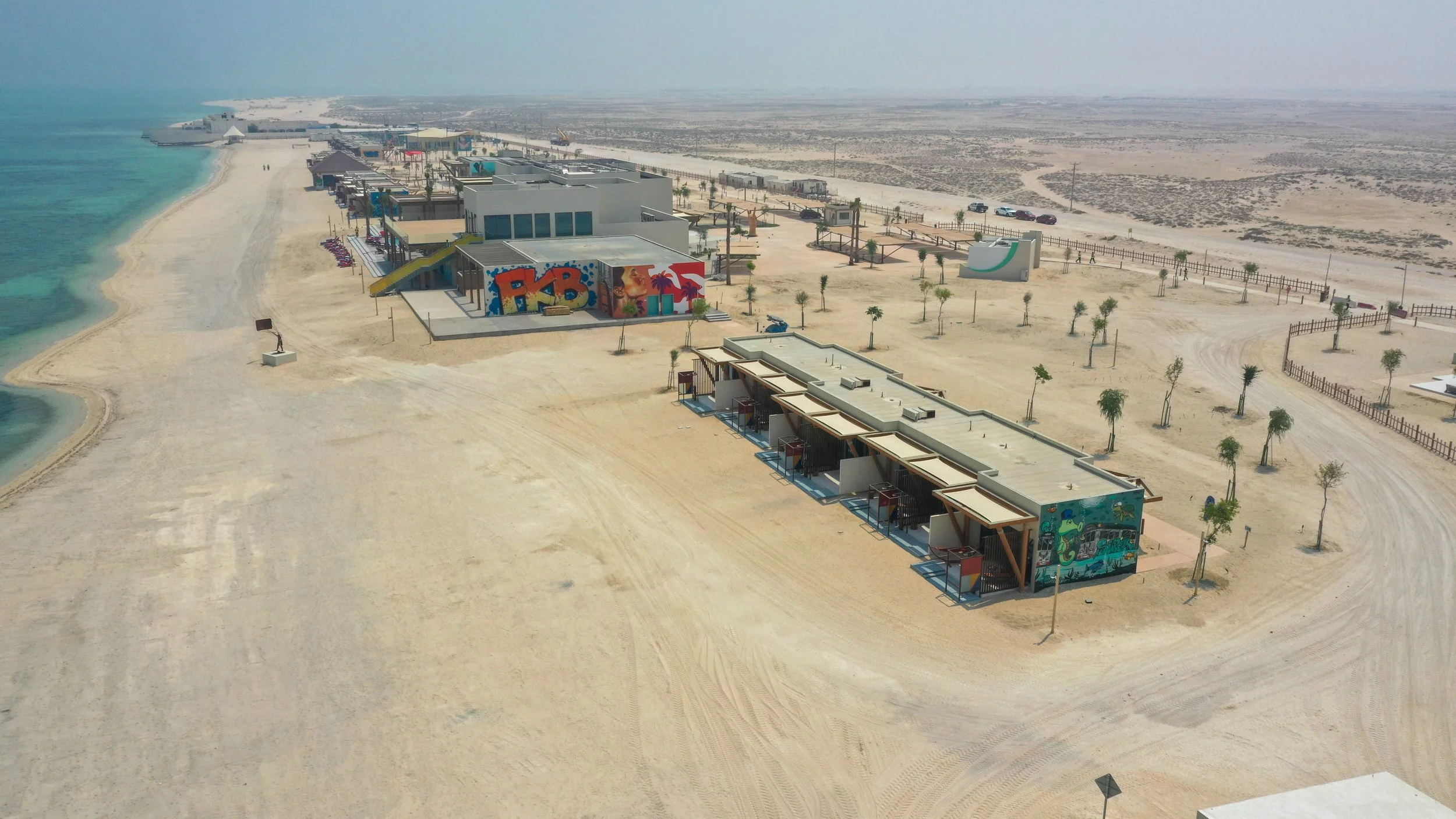 Aerial view of an empty beach resort with colorful graffiti art, modern buildings, palm trees, a sandy beach, and a distant road in a desert-like landscape.