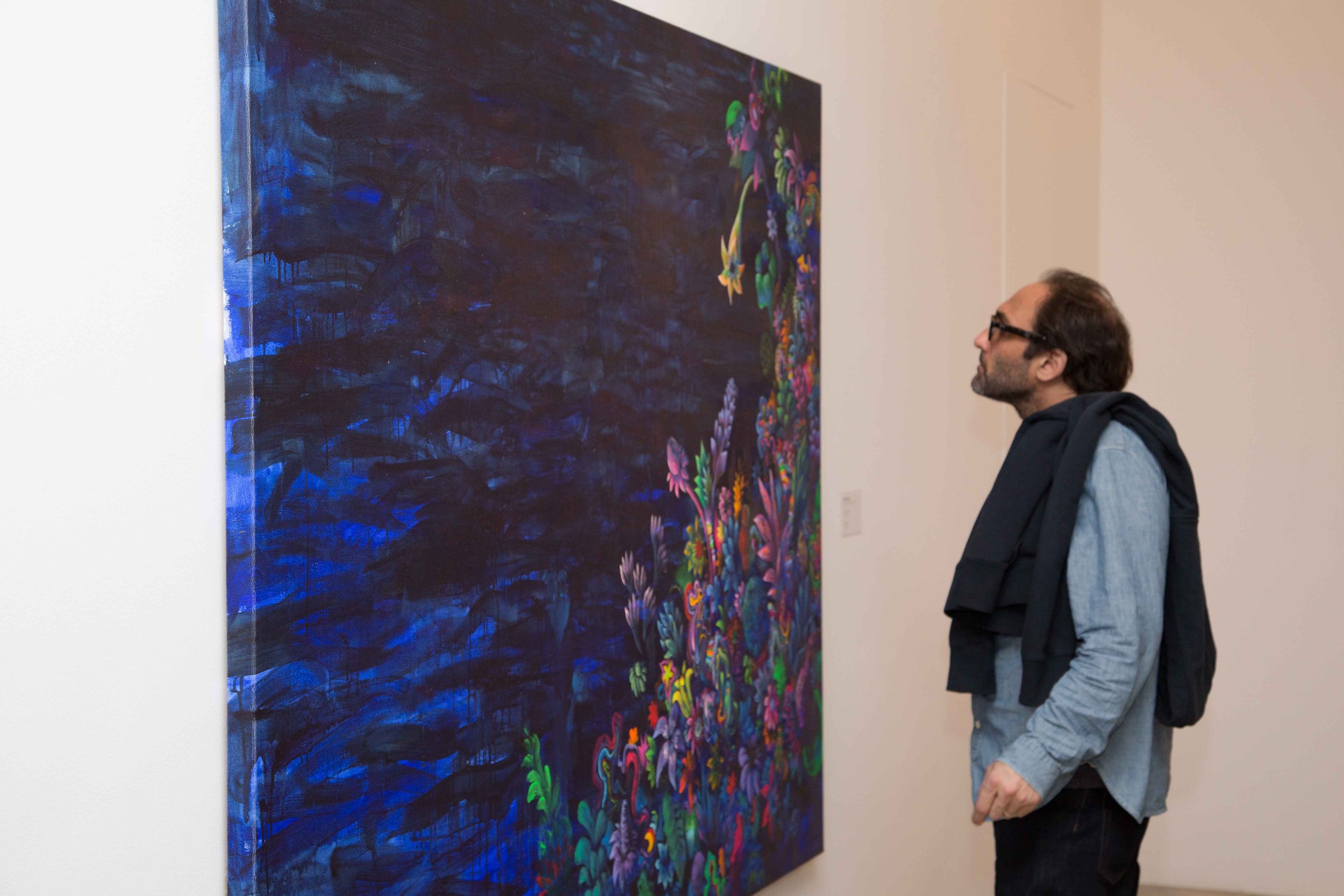 A man in glasses looking at a large abstract painting in an art gallery. The painting features dark blue background with colorful, vibrant floral and organic shapes.