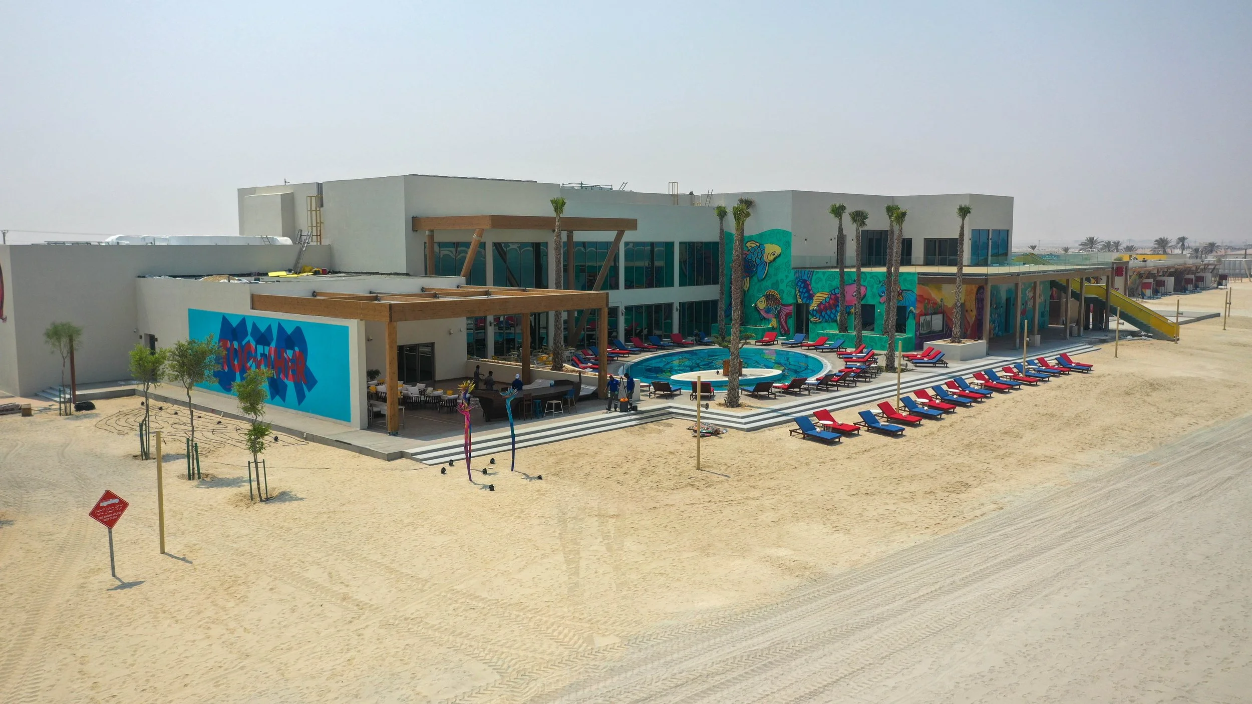 A beachfront resort with colorful murals, a pool, and red and blue lounge chairs on sandy terrain.