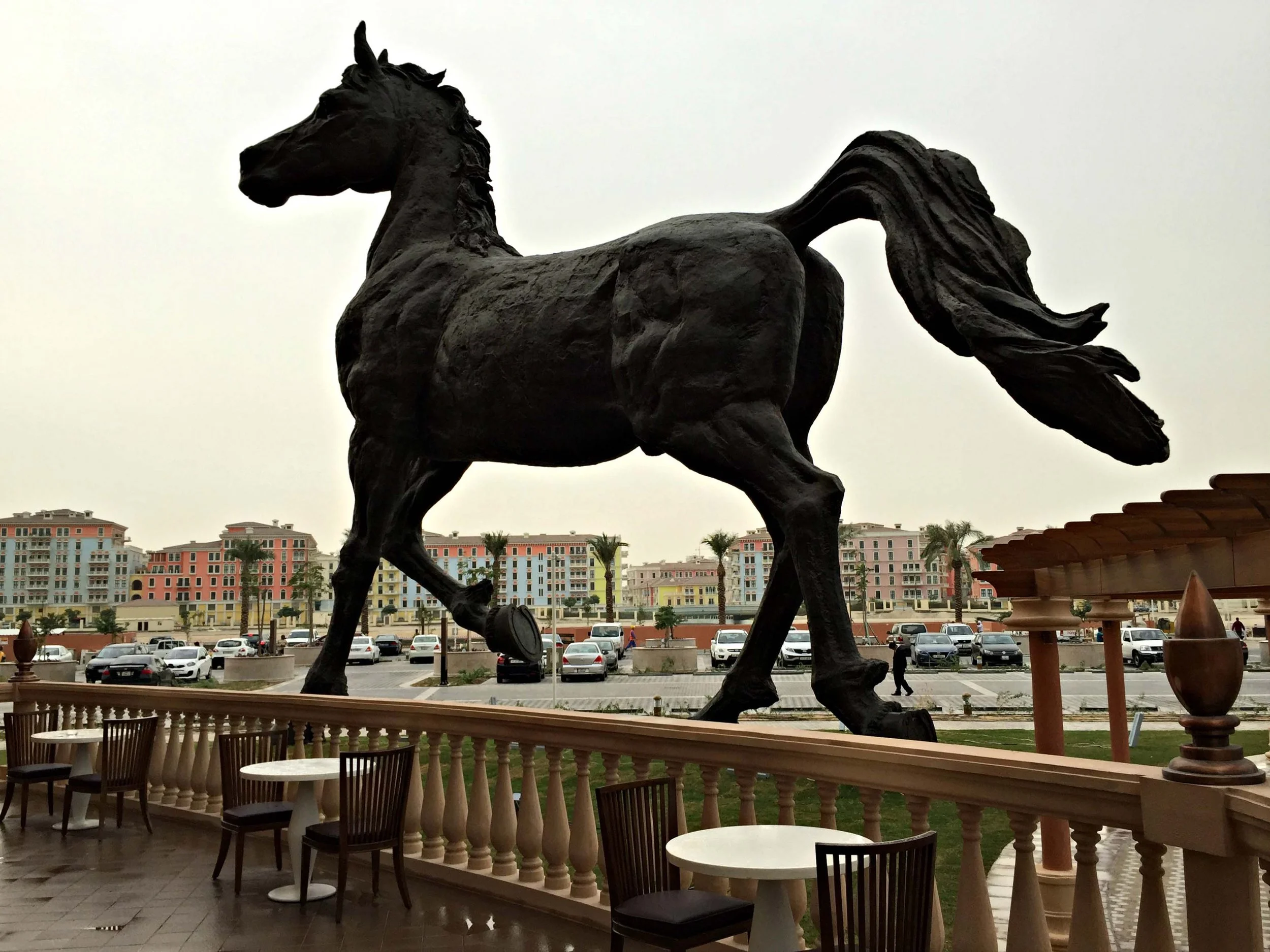 Large black statue of a running horse outside a building, with a parking lot and colorful apartment buildings in the background.