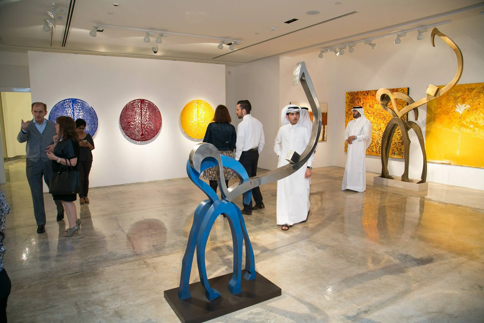 People touring an art gallery with colorful paintings and sculptures, including a blue and silver abstract sculpture in the foreground.