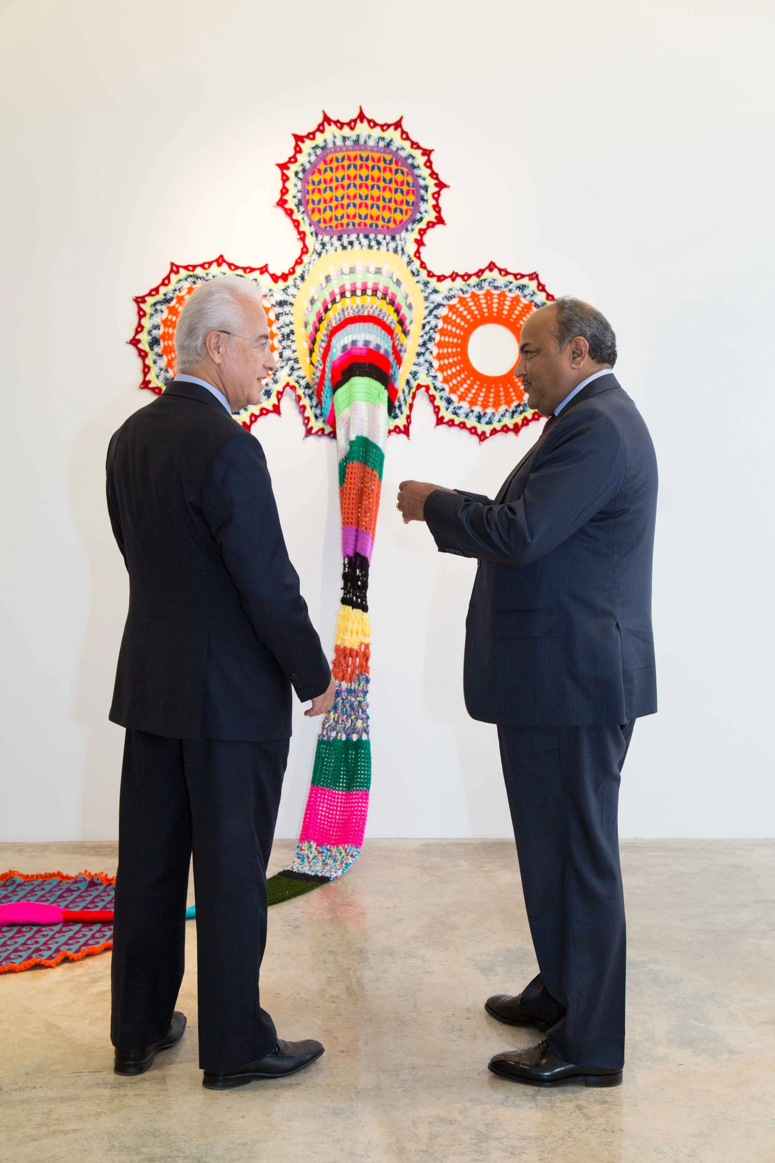 Two men in suits standing and talking in front of a colorful, abstract yarn artwork on a white wall.