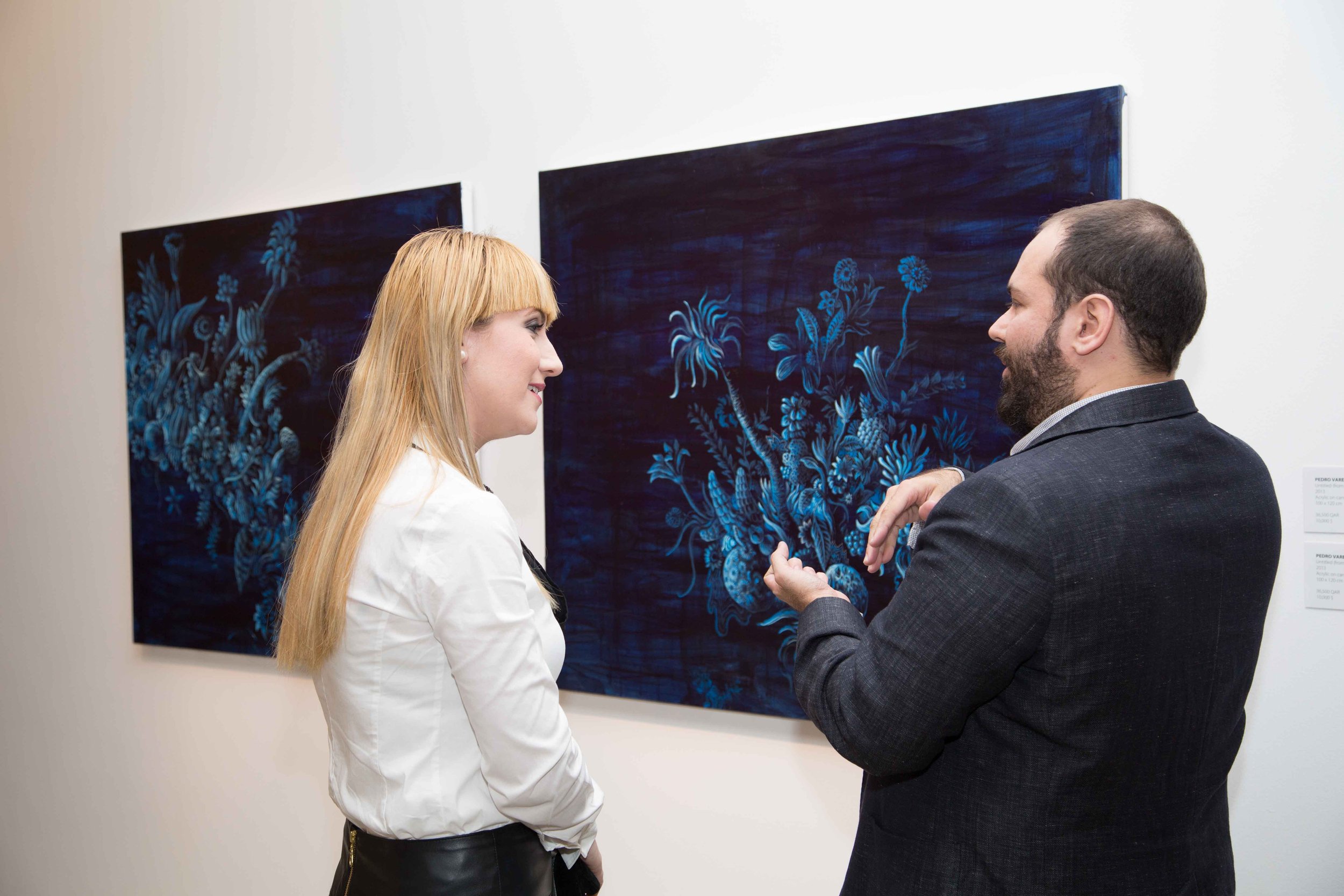 A man and woman having a conversation in an art gallery, with blue floral paintings on the wall behind them.