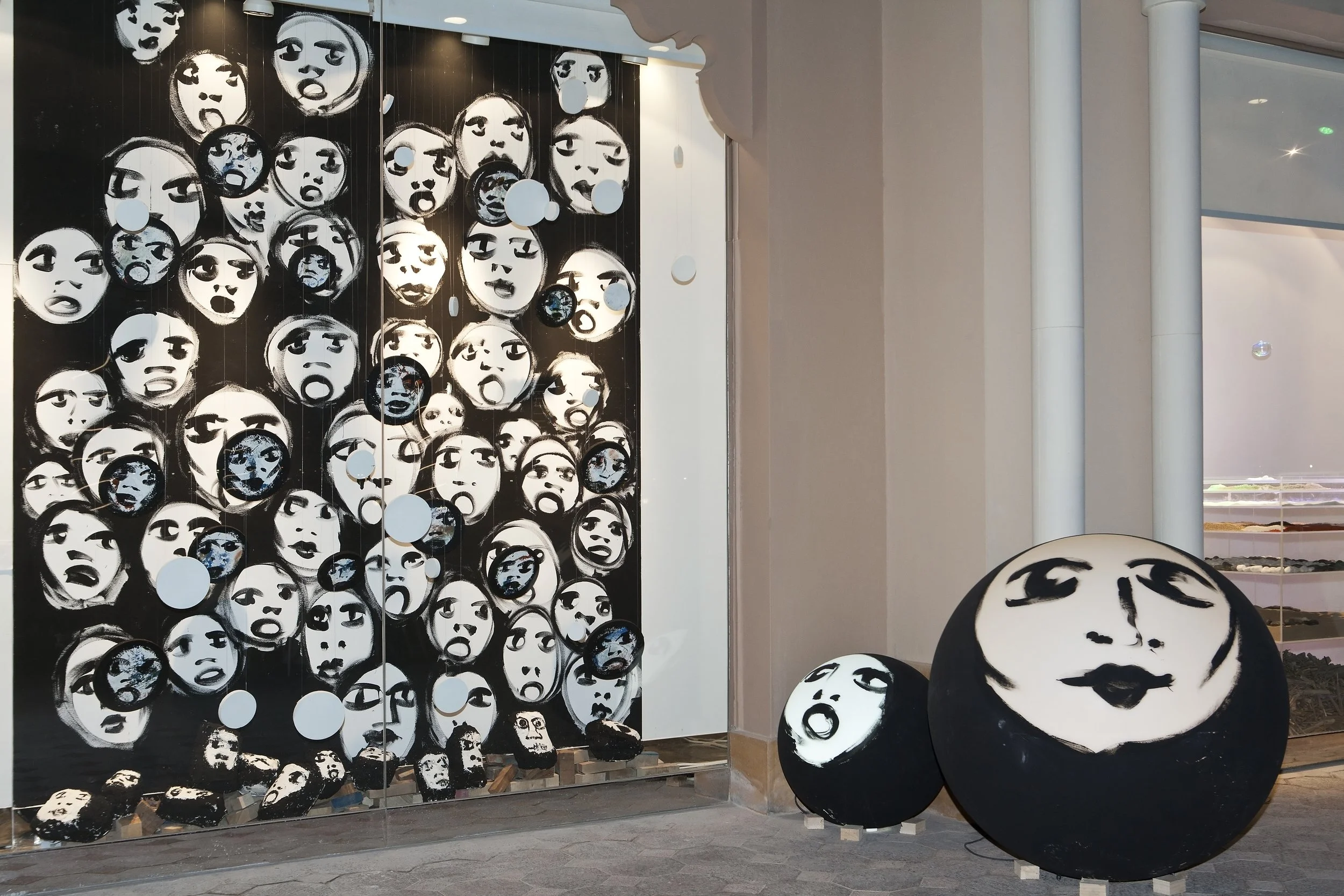 Outdoor art installation featuring a large wall of black-and-white painted faces with various expressions, along with three nearest large round sculptures also painted with face designs, set against a neutral wall and a window display in the backgrou