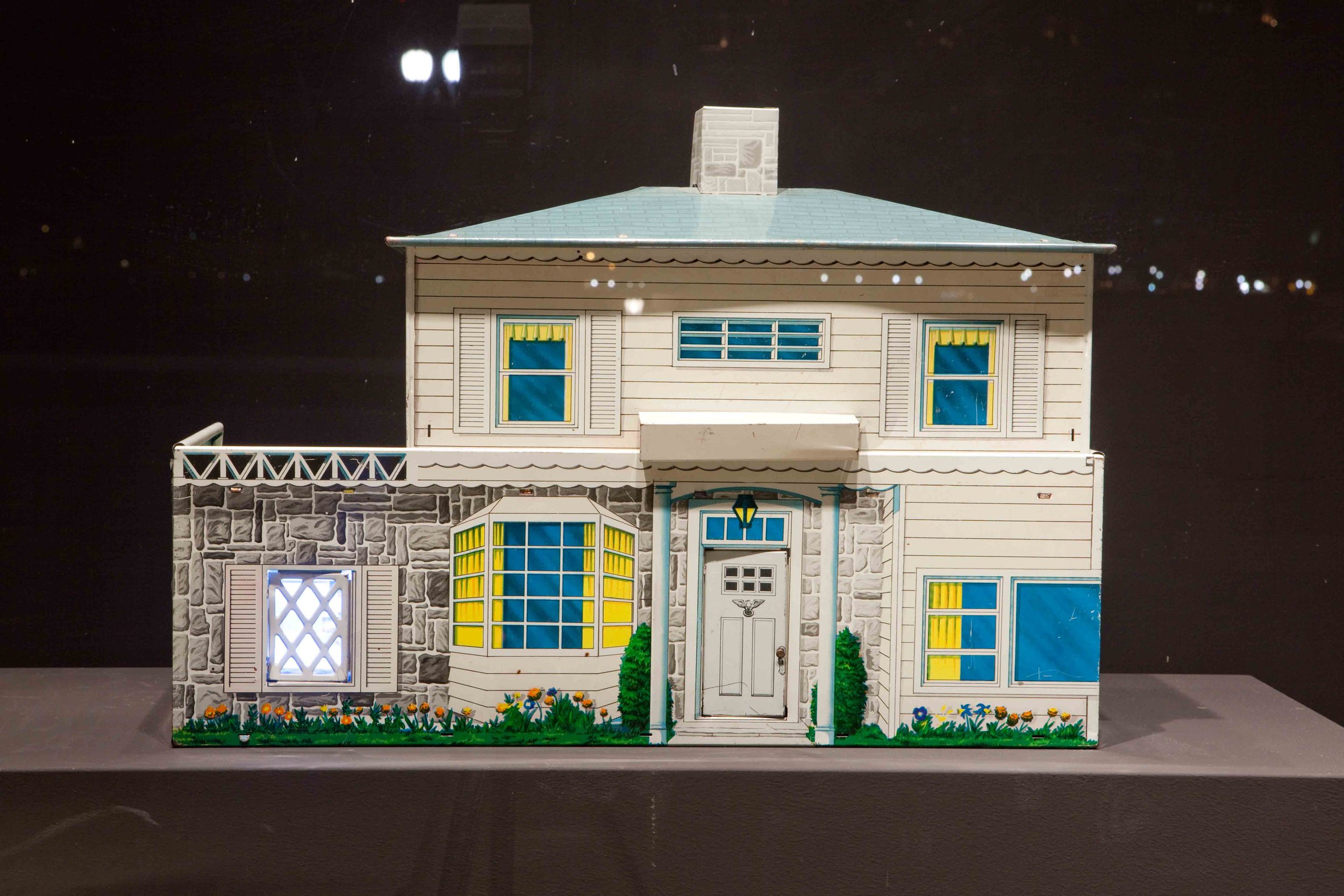 A miniature model of a two-story house with stone and siding exterior, multiple windows with yellow curtains, a front door with a lantern, and a small garden with flowers in front.