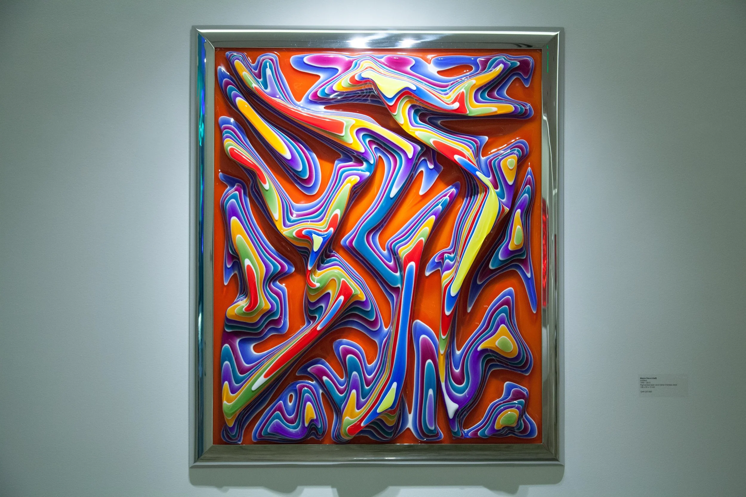 Abstract colorful swirl art piece with wavy lines on an orange background, framed and displayed on a gallery wall.