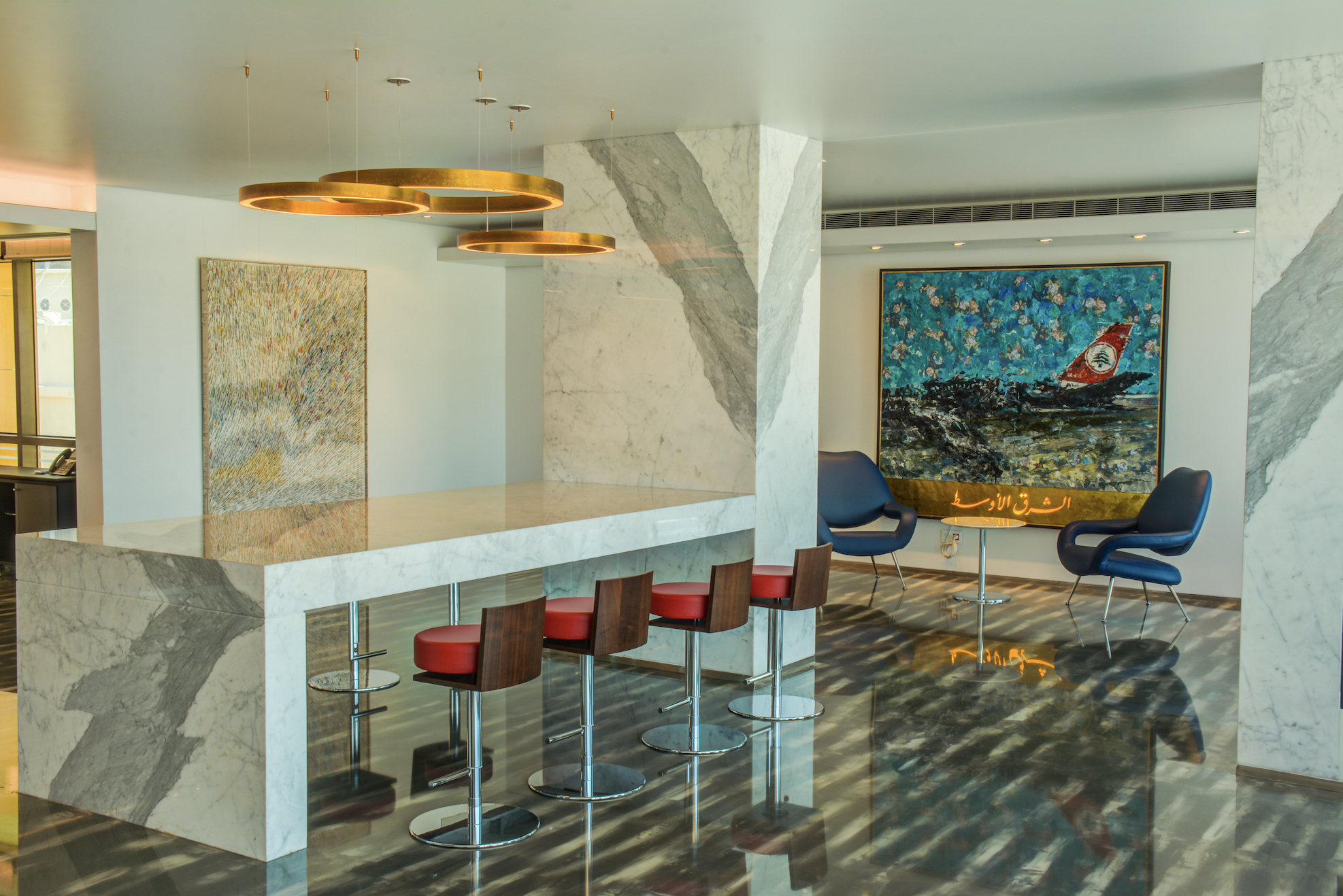 Modern hotel lobby area with a marble bar counter, four red bar stools, two blue lounge chairs, and a large abstract painting with Arabic writing on a gold plaque, set on a reflective black floor.
