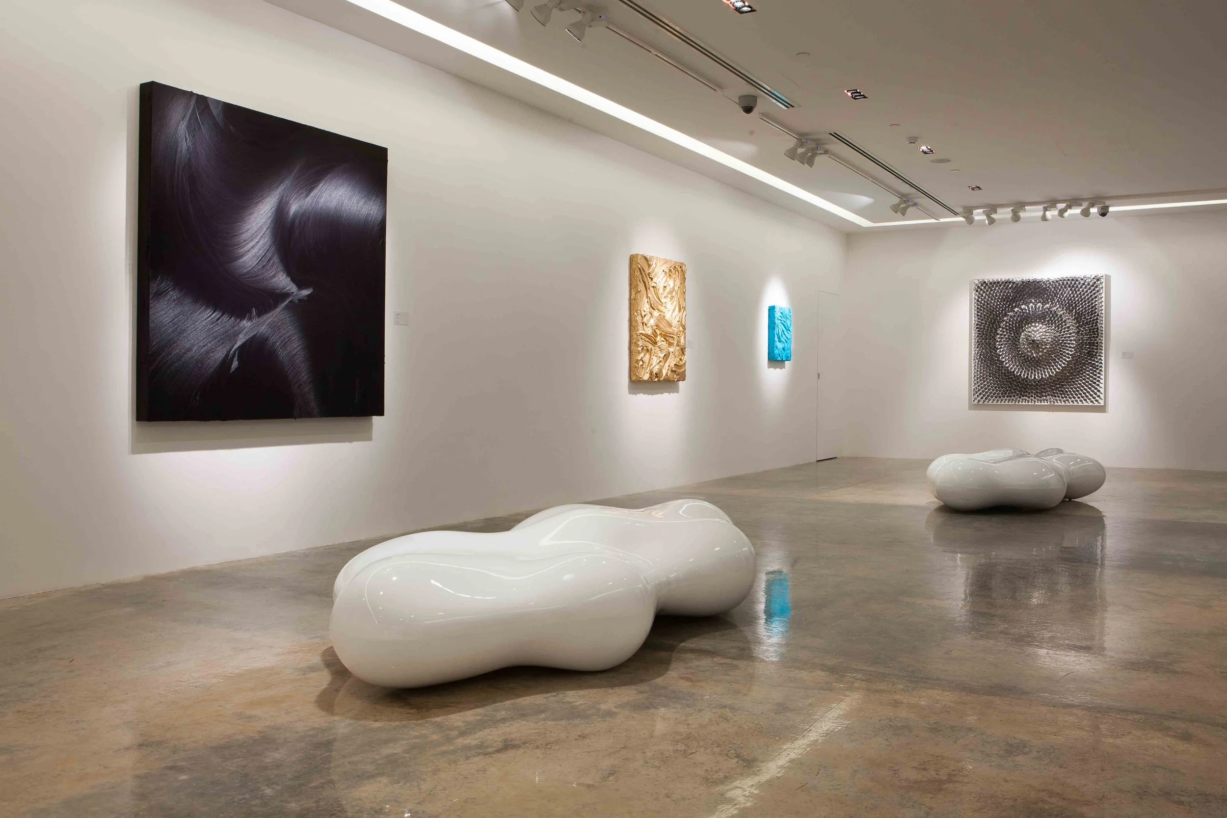 An art gallery with white walls displaying five artworks: a large black abstract painting, a textured gold art piece, a blue textured art piece, a geometric black and white piece, and a black textured art piece. Two glossy white sculptural benches wi