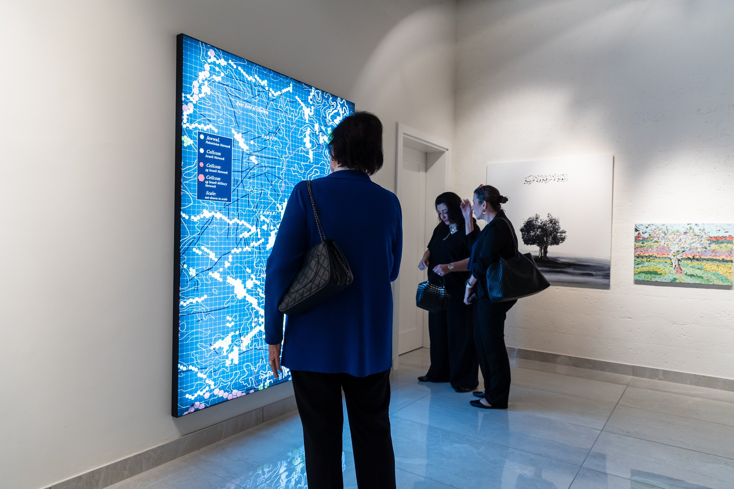 Four women in black and blue clothing are viewing art and digital displays in an art gallery, with a large digital map and several paintings on white walls.