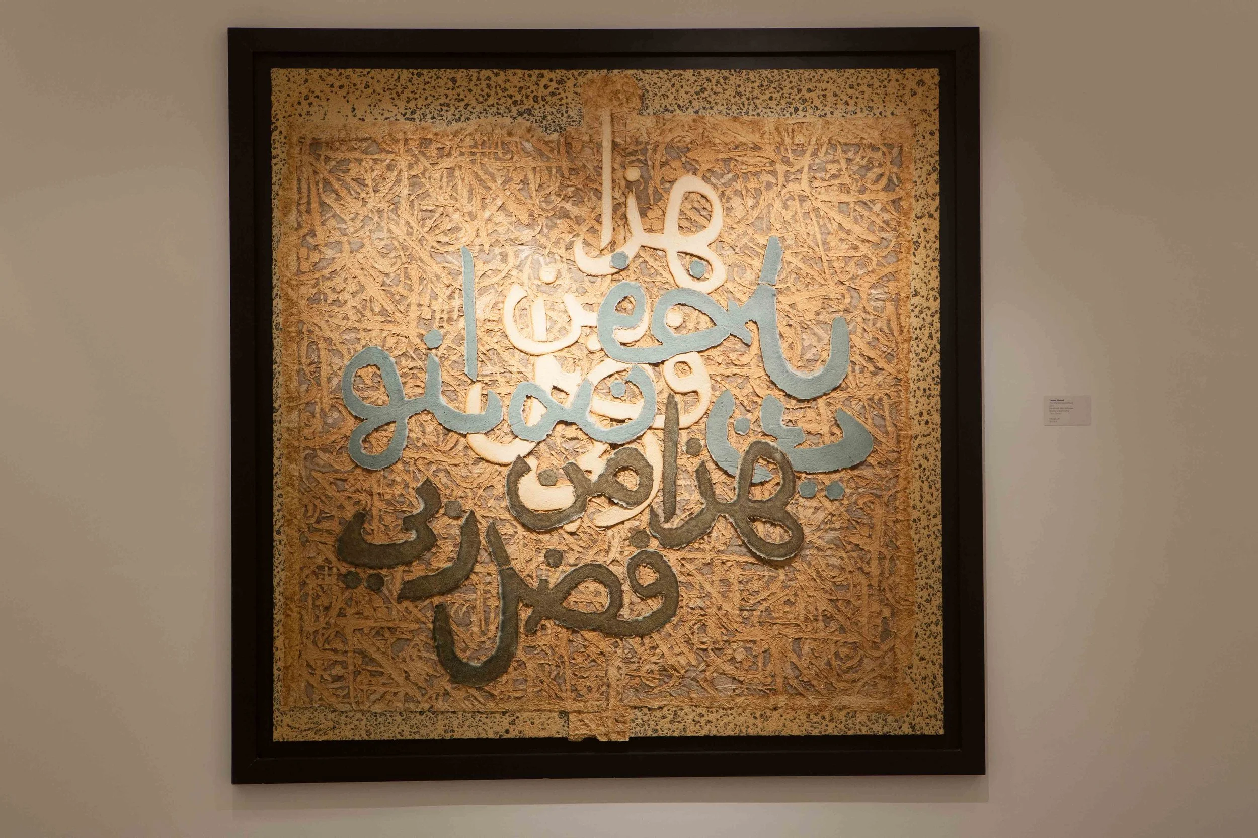 Framed artwork featuring Arabic calligraphy with metallic silver and gold accents on a textured beige background.