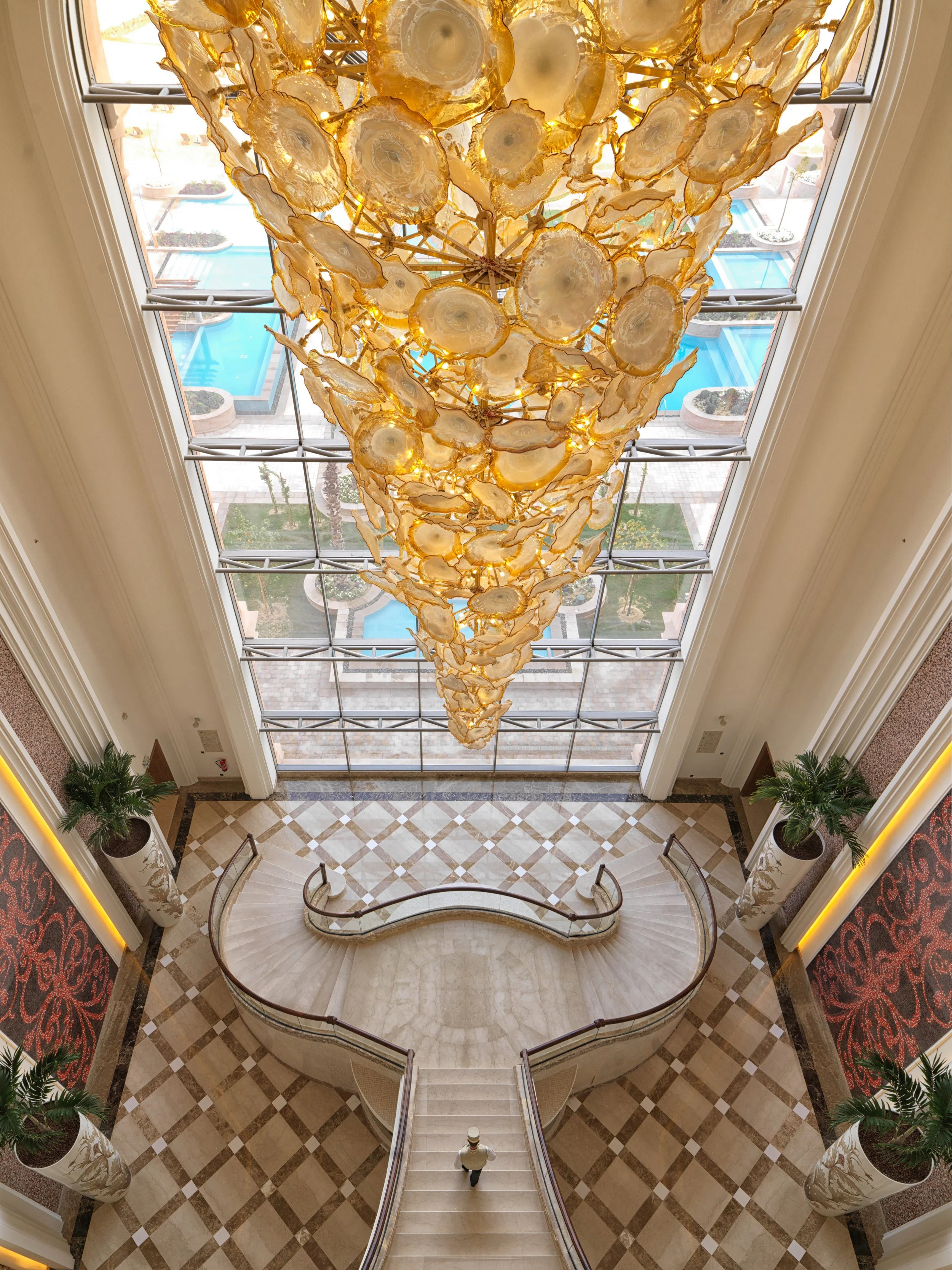 A grand hotel lobby with a large, ornate chandelier hanging from the ceiling, viewed from above. The chandelier is decorated with numerous slices of agate and warm lighting. Below is a curved staircase with a single person walking down, flanked by la