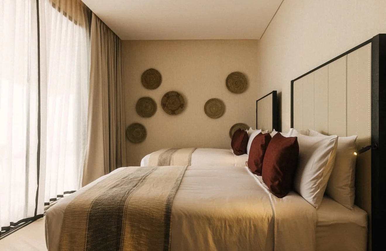 Hotel room with two large beds, beige headboards, and brown pillows. Wall art with circular woven designs, cream curtains, and soft lighting.