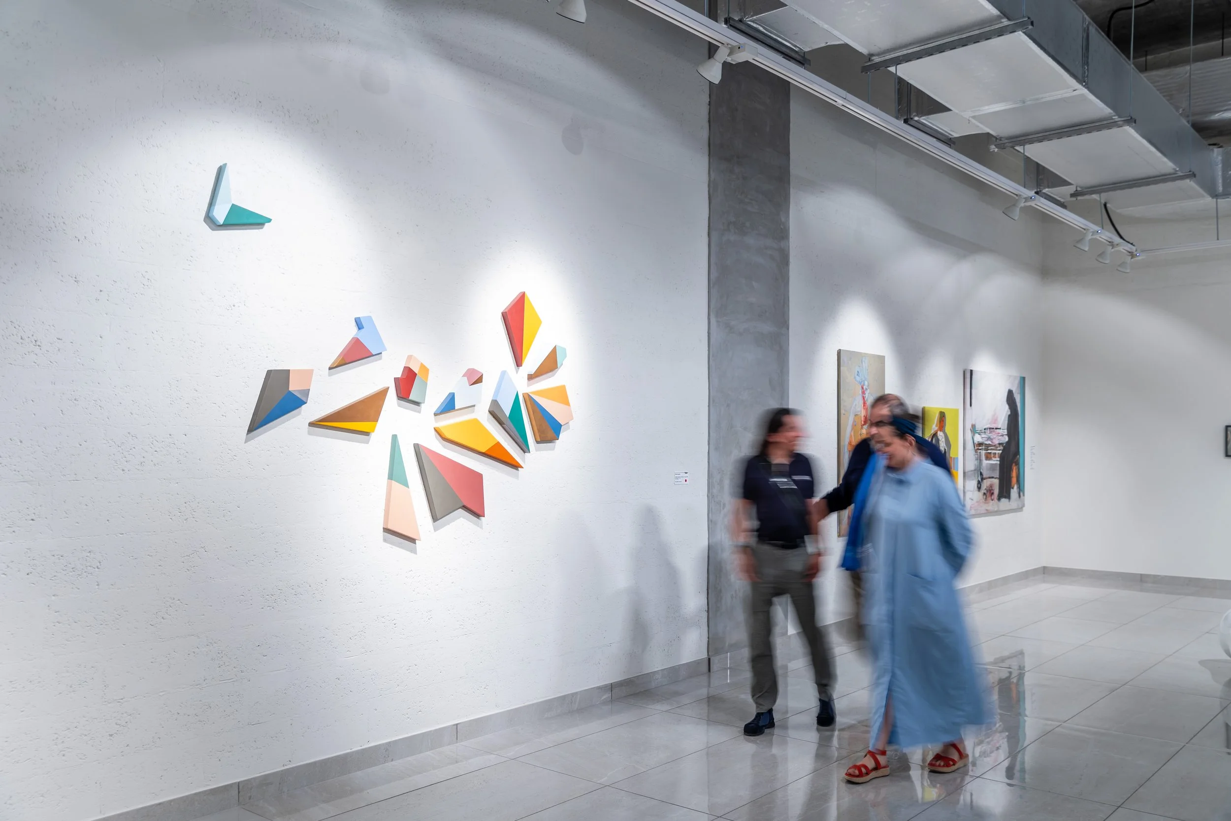People viewing colorful abstract art on gallery wall with plain white and concrete walls and tiled floor.