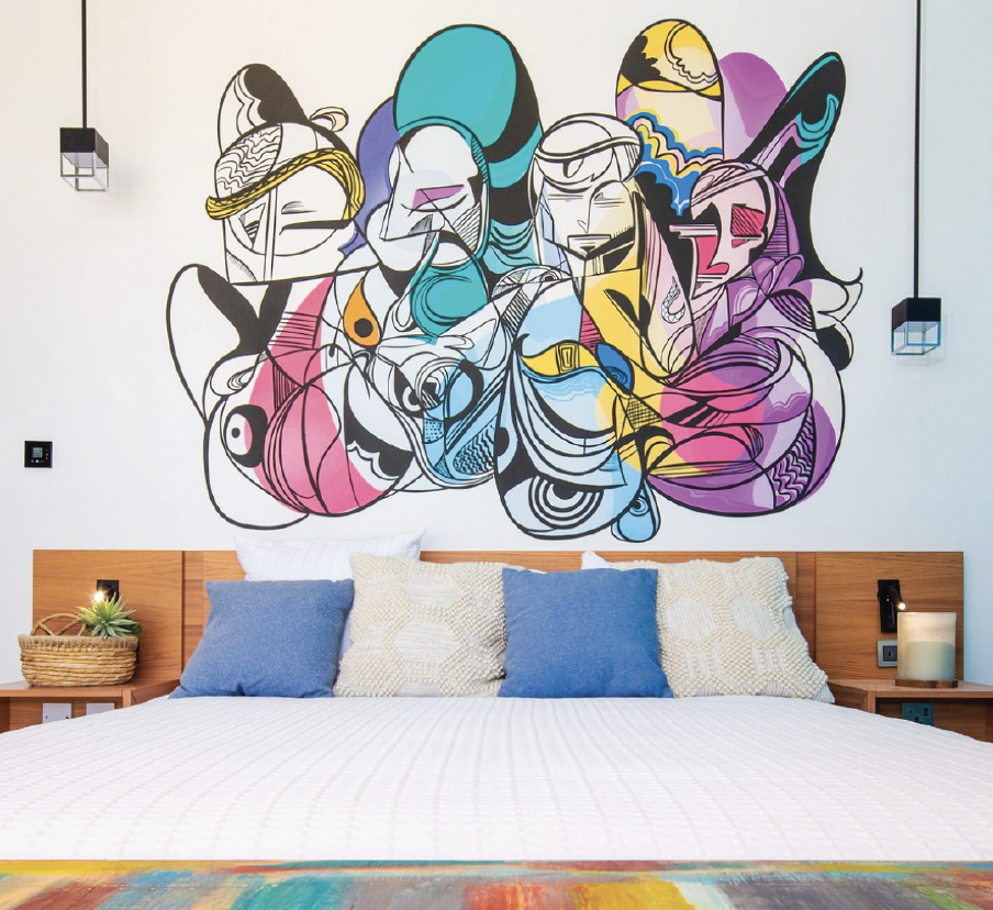 Colorful wall art above a bed in a bedroom, with decorative pillows, a bedside table with a plant, and a painted rug at the foot of the bed.