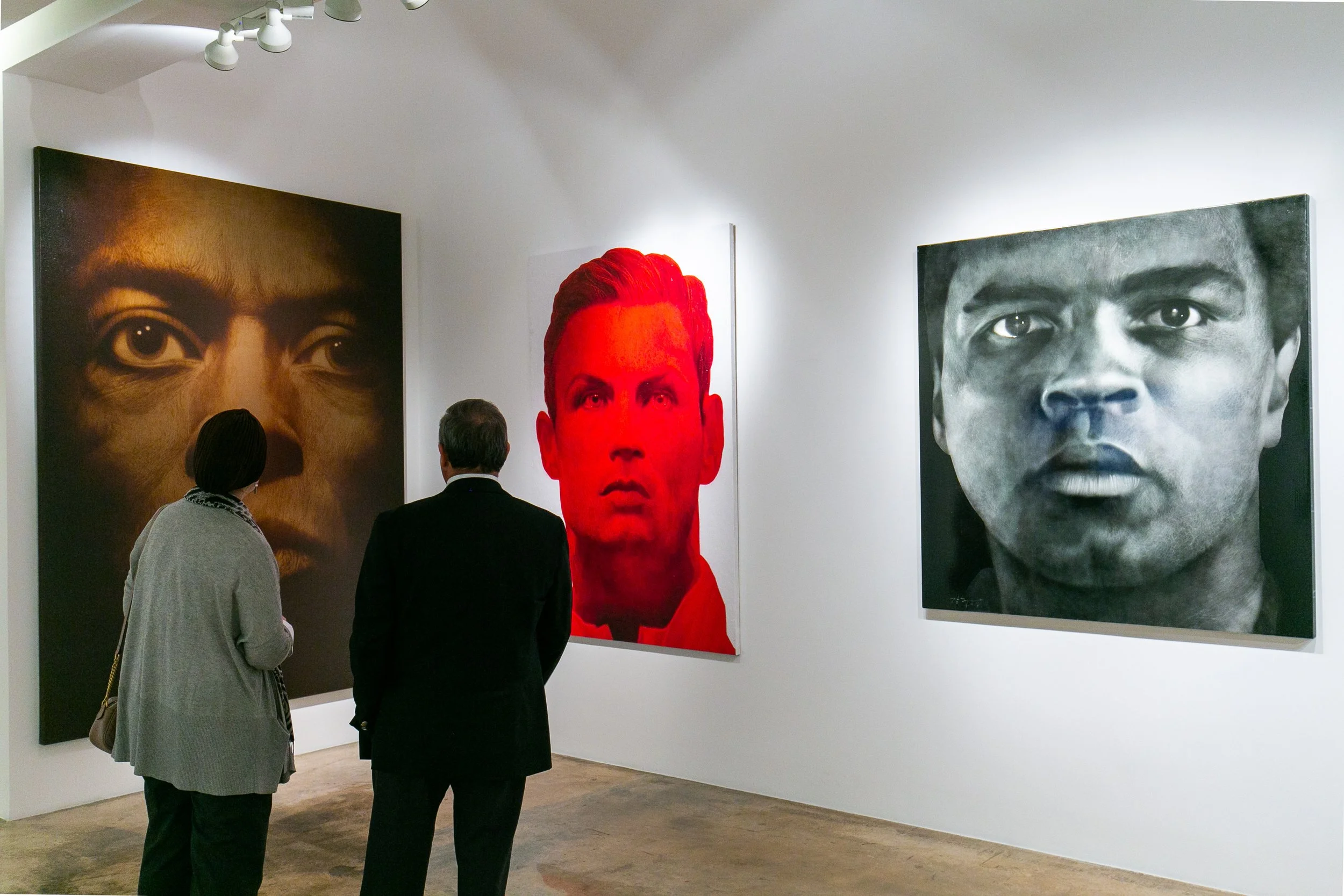 Two people observe large black-and-white and color portraits of diverse individuals in an art gallery.
