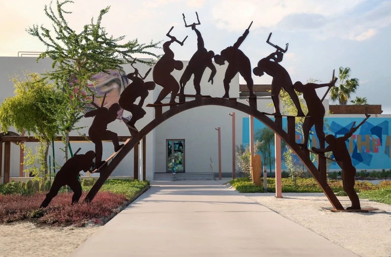 Metal sculpture of muscular figures carrying a beam over an arch, representing teamwork and strength, situated in an outdoor park with trees and a building in the background.