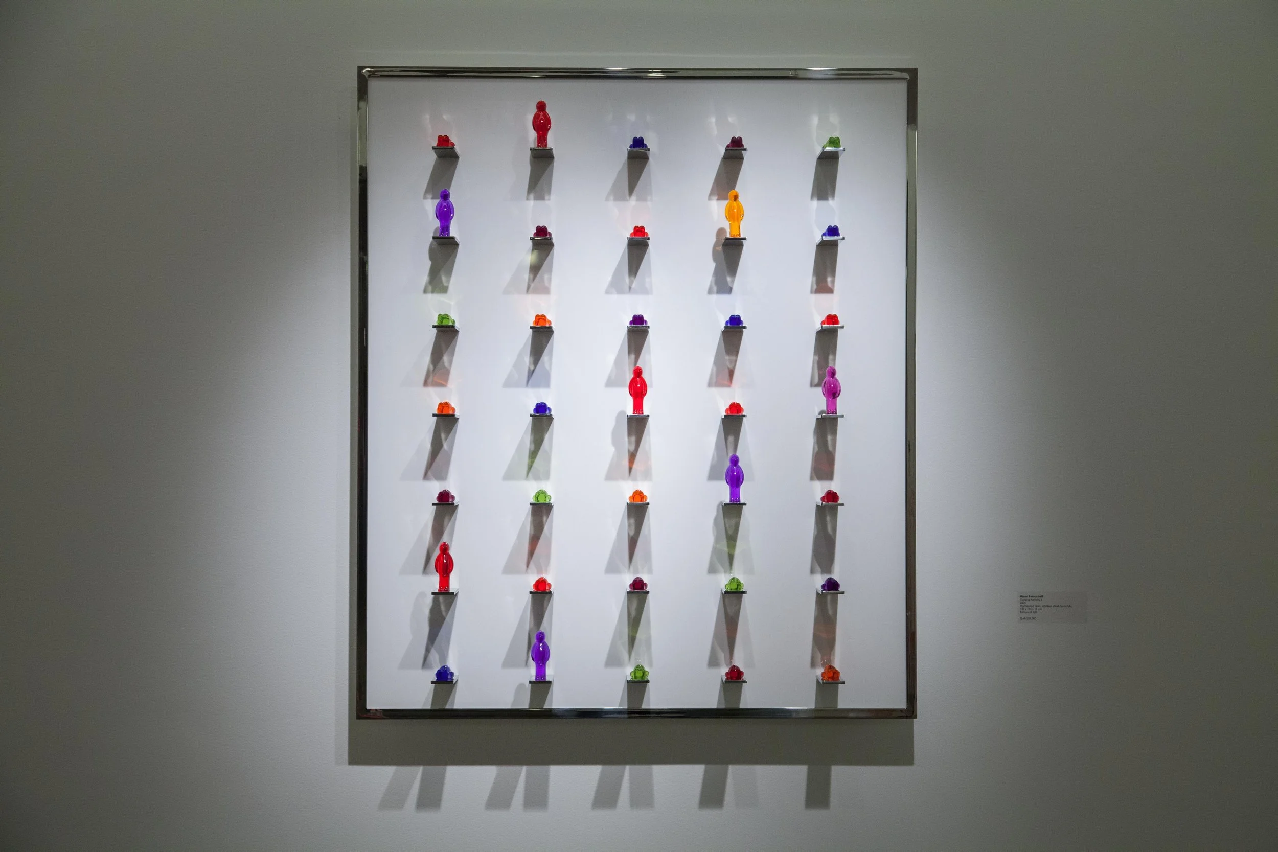 Colorful miniature figurines arranged on black shelves in a grid on white background, displayed as a contemporary art piece.