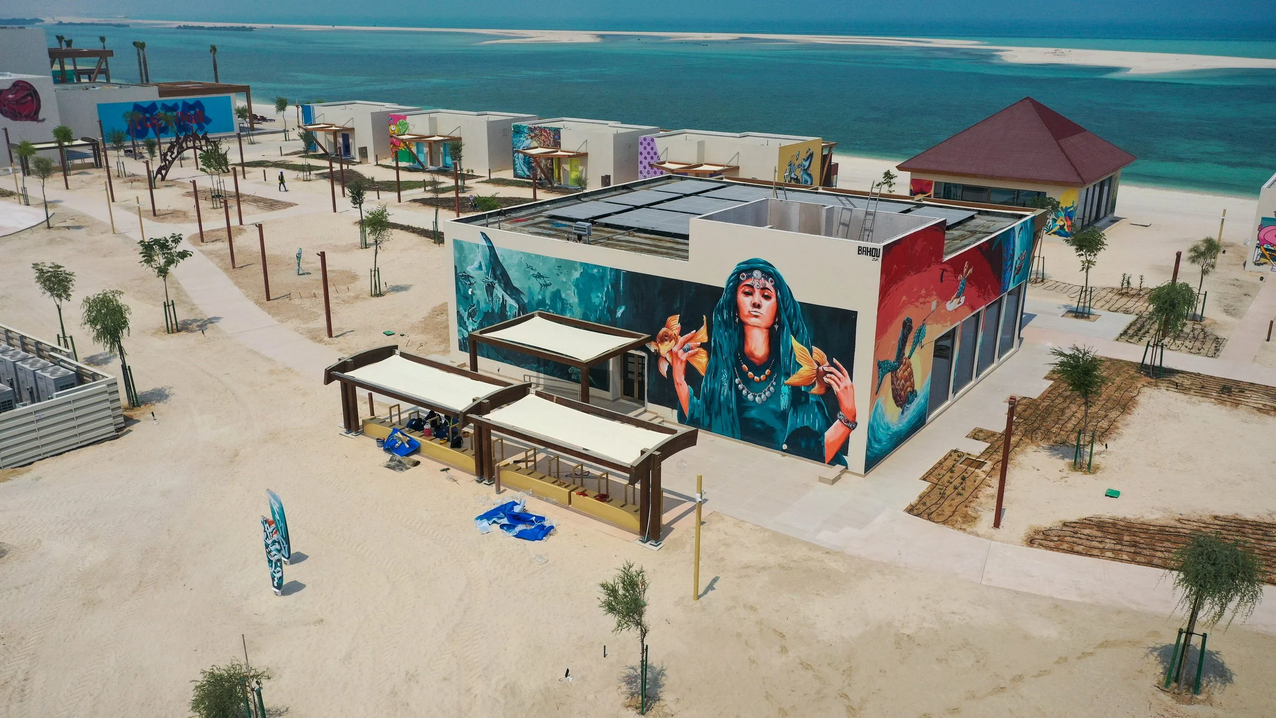 An aerial view of an art installation on a beachfront with graffiti murals, small trees, and sandy walkways.
