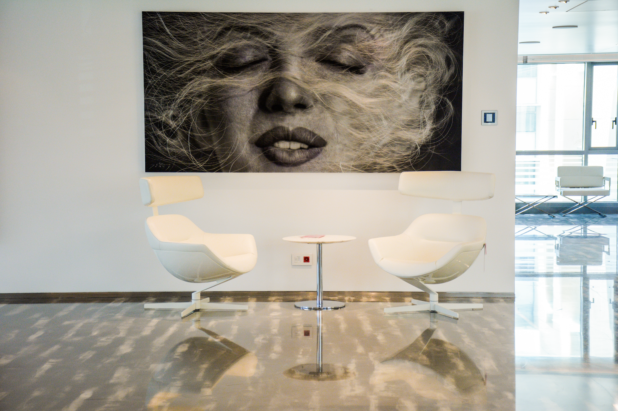 Modern lounge area with two white chairs and a small round table between them, with abstract black and white portrait artwork of a woman with flowing hair on the wall behind.