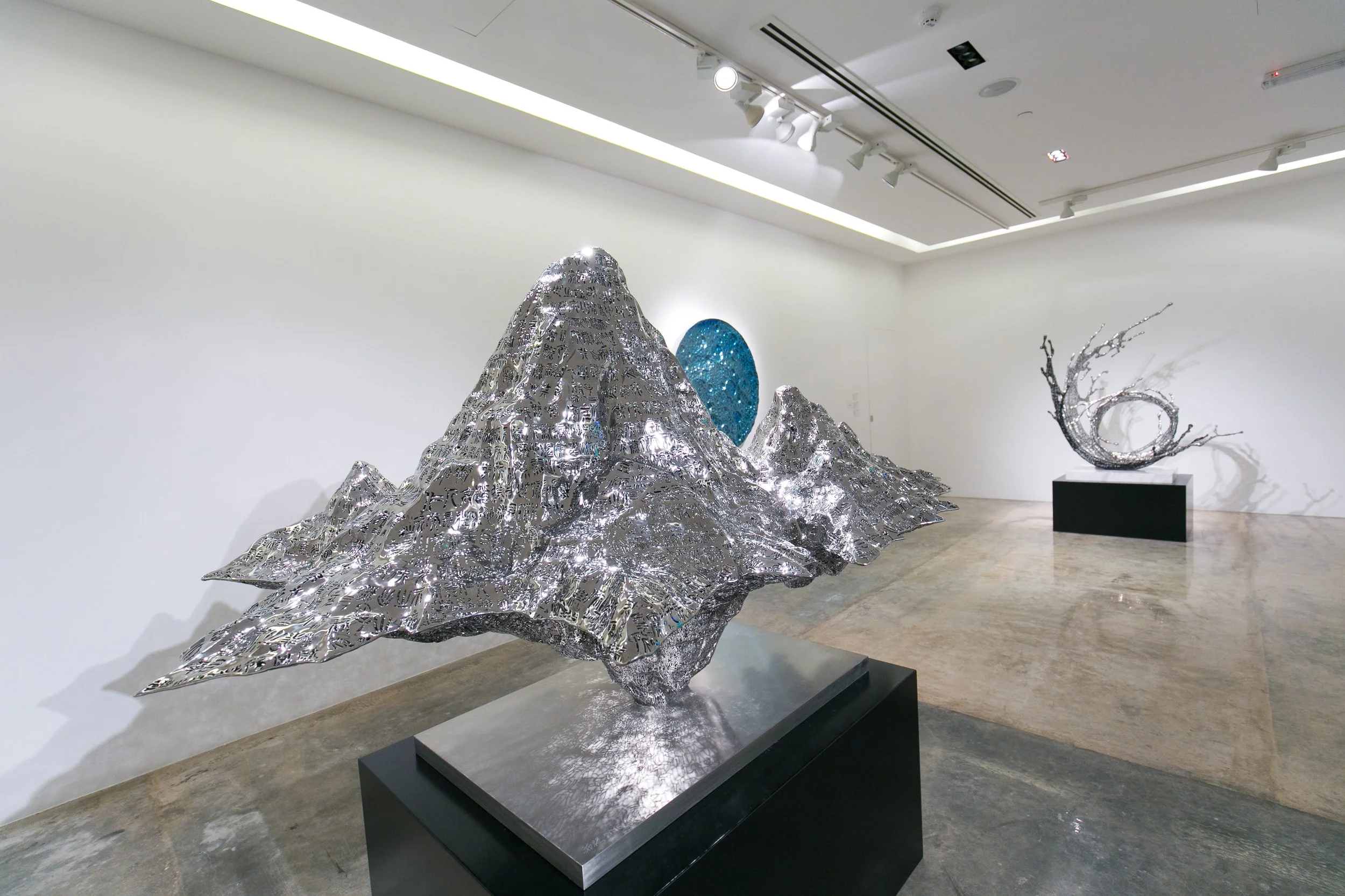 Art sculptures in a gallery, including a mountain-shaped metallic sculpture in the foreground and a crescent-shaped sculpture on the right side with branch-like extensions.