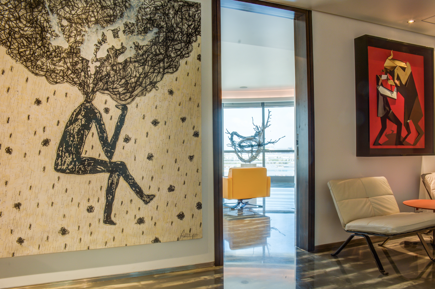 Interior view of a modern art gallery with colorful paintings on the wall. A large black and white artwork on the left shows a figure sitting with a cloud of tangled lines above its head. Through a doorway, a seating area with a yellow chair and an a