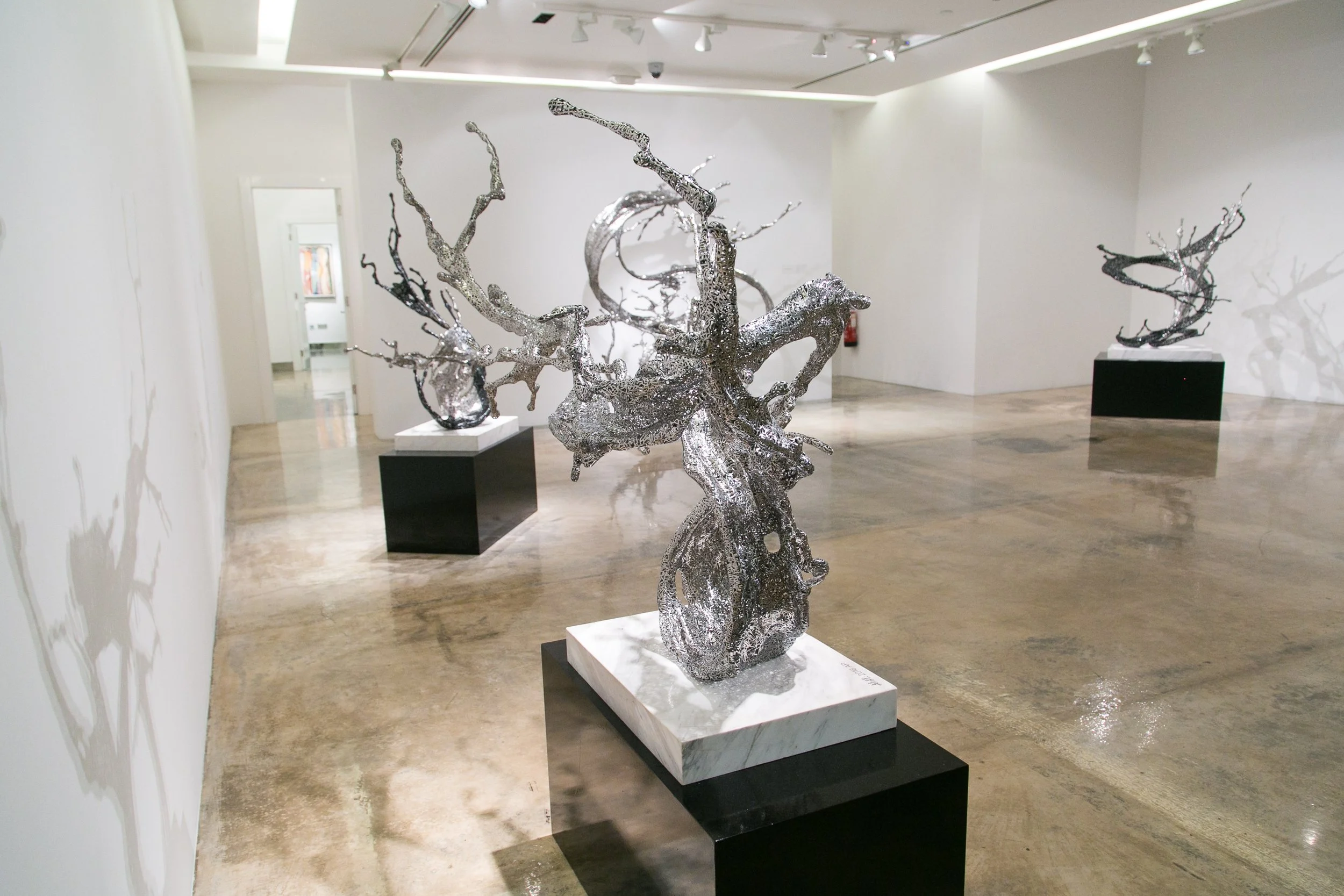 A shiny metallic sculpture of a twisted tree on a white and black pedestal in an art gallery with white walls and a polished concrete floor.