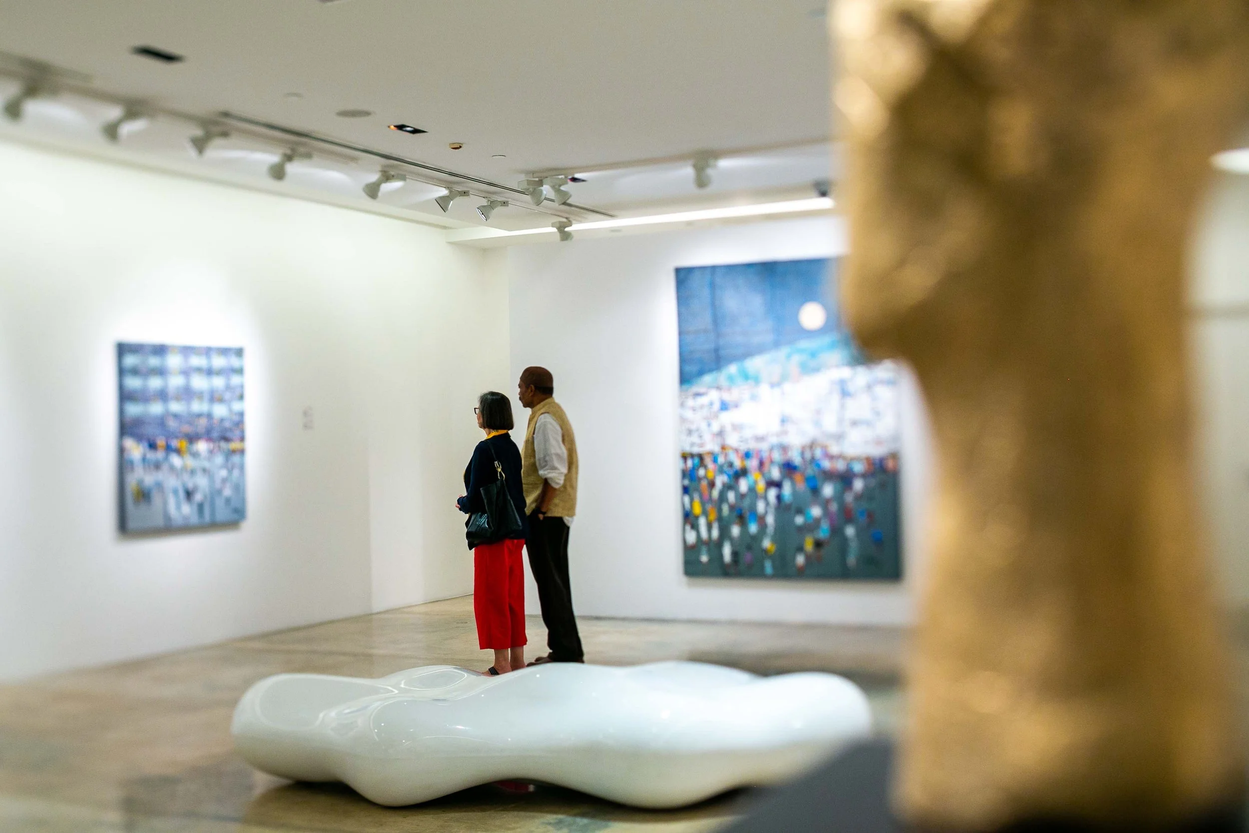 Two people stand in an art gallery looking at paintings on white walls, with a blurred sculpture in the foreground.