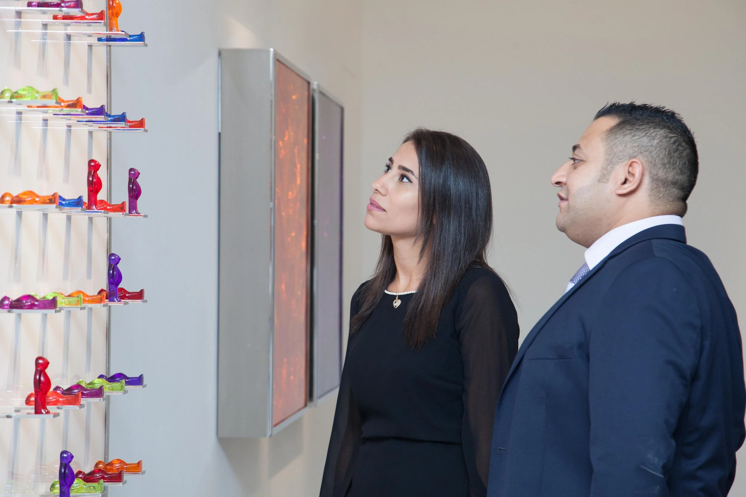 A man and woman in business attire observing colorful figurines on a wall display.