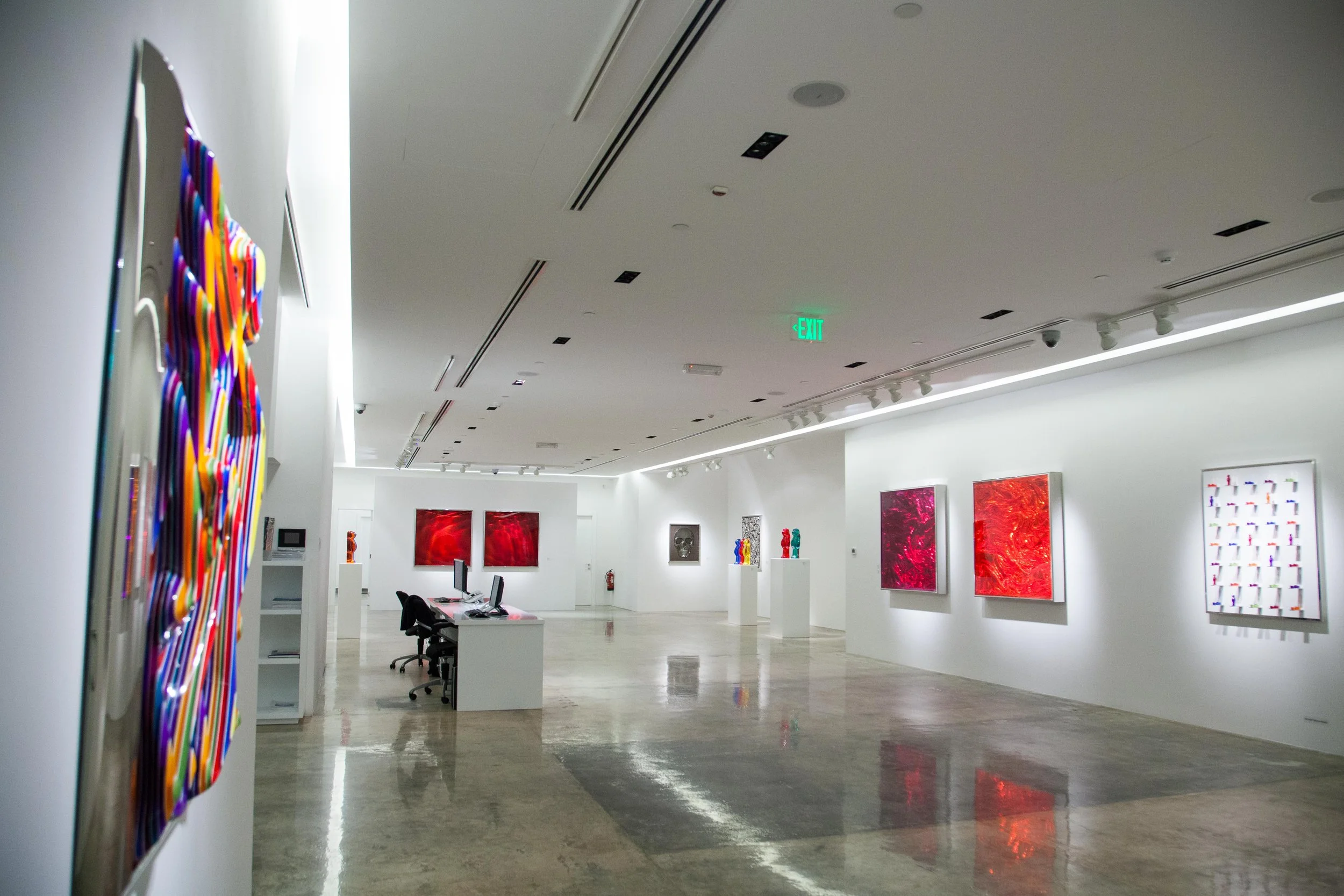 An art gallery with white walls displaying colorful paintings and sculptures, with a polished concrete floor and modern lighting.