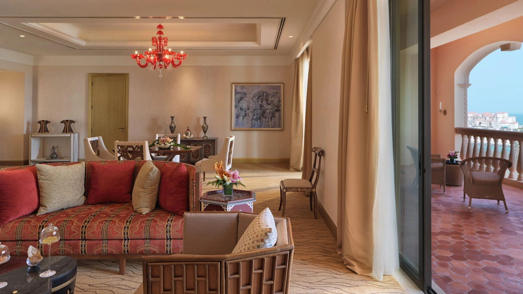 Spacious hotel living room with red chandelier, cream walls, and sliding glass door leading to balcony with outdoor seating and ocean view.