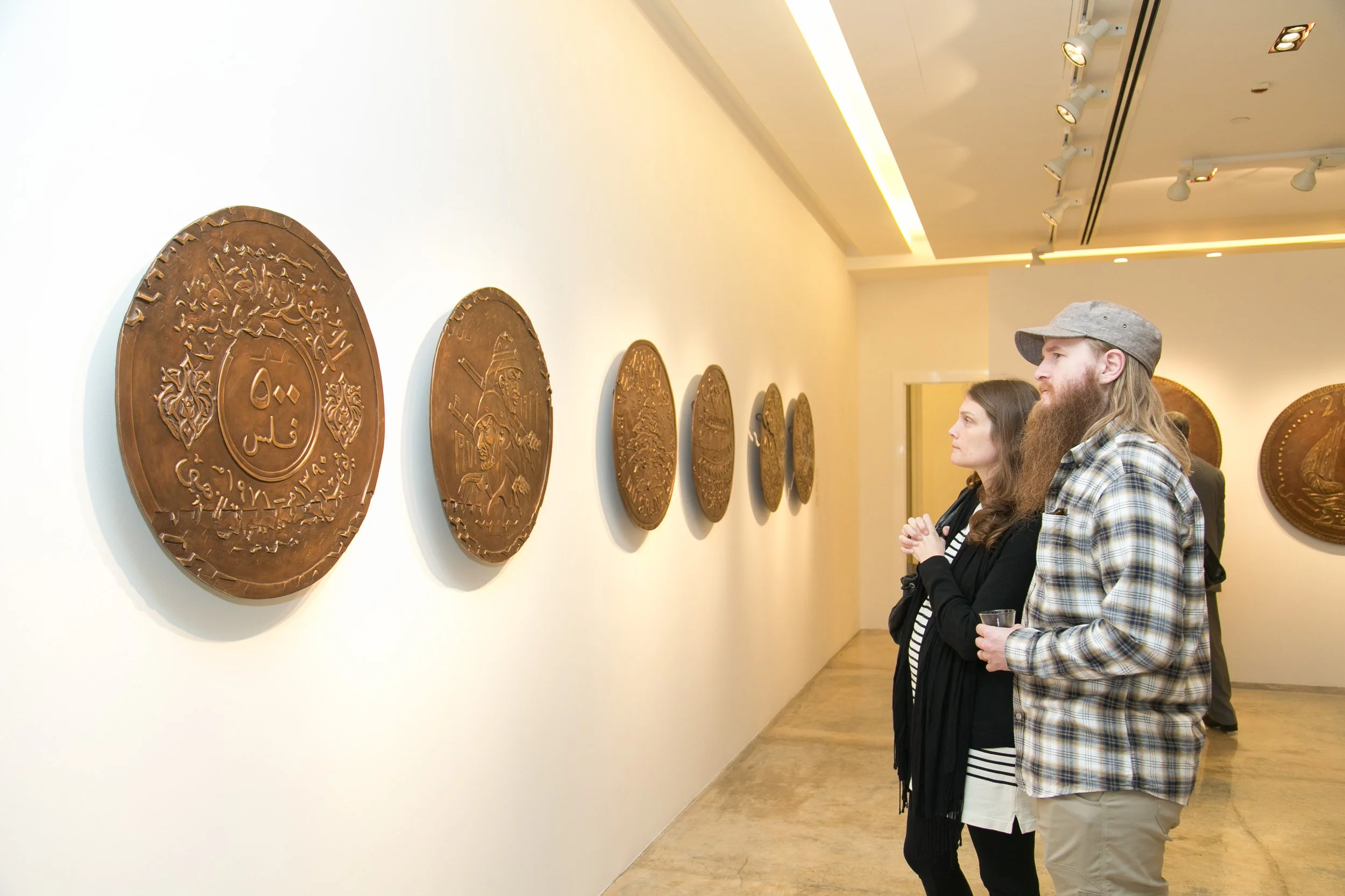 Two people, a man with a beard in a baseball cap and plaid shirt, and a woman with long brown hair in a black jacket, observe bronze coins displayed on a white museum wall.