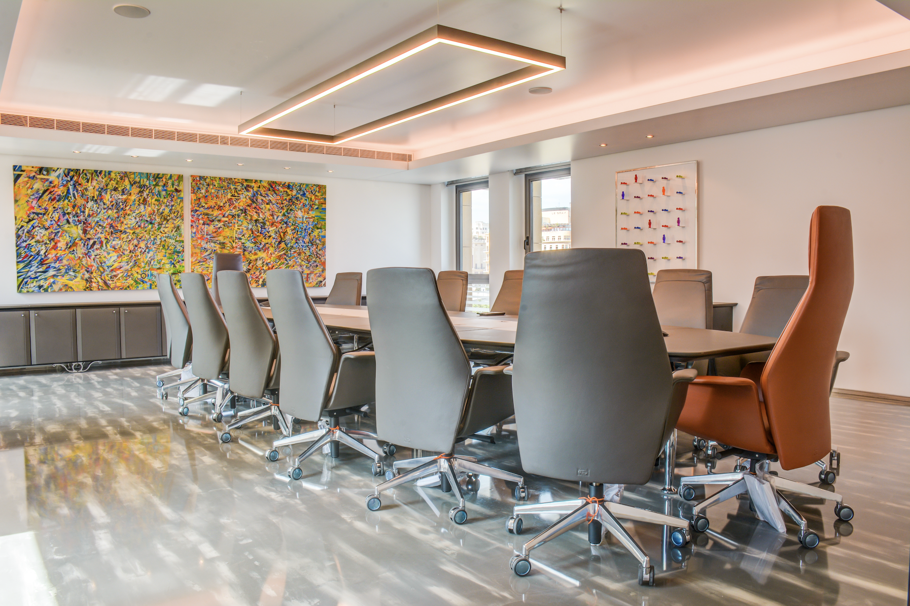 Modern conference room with a long table surrounded by high-backed office chairs in gray and brown. Large colorful abstract artwork on the wall, with natural light coming through windows.