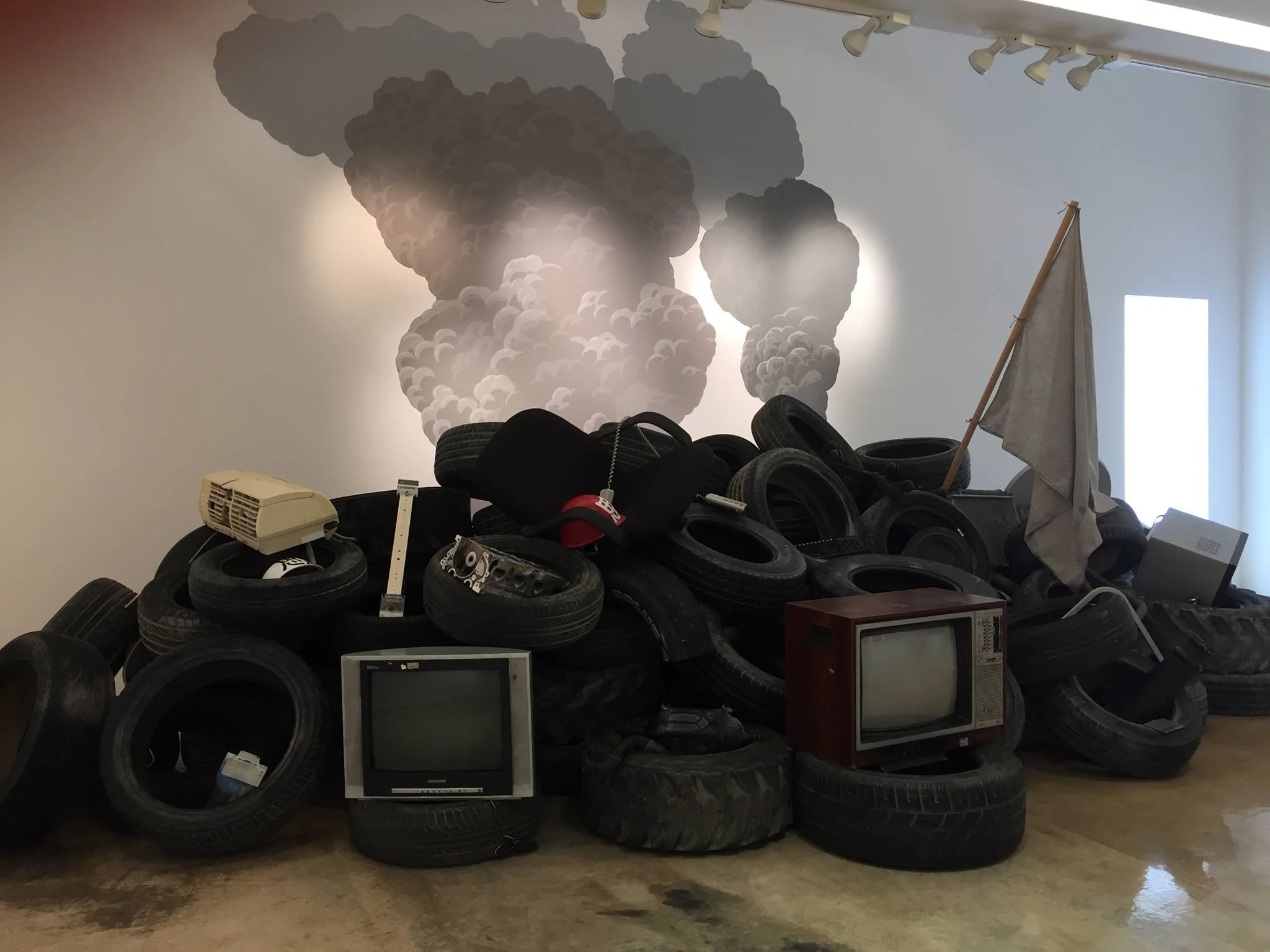 An art installation depicting a pile of old tires, vintage televisions, and various objects with a cloud of gray smoke painted on the wall behind, and a white flag on a stick among the debris.