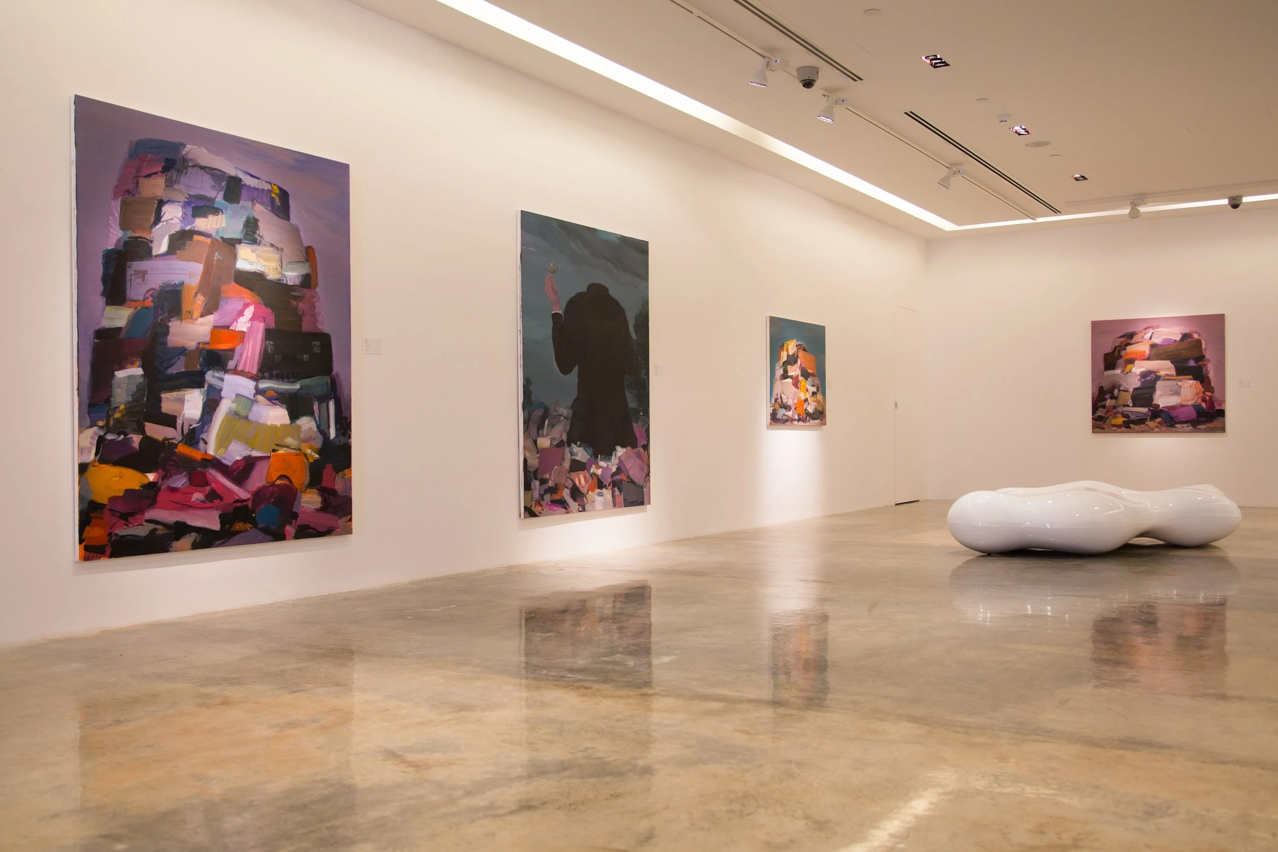 An art gallery with white walls and polished concrete floors displaying five abstract and figurative paintings, some with colorful brushstrokes, and a sleek white modern bench in the center.
