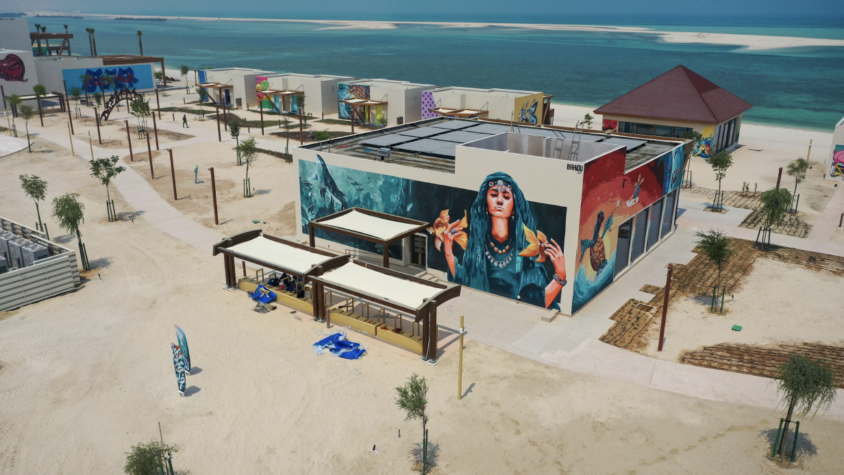 An aerial view of a beachside area with colorful murals on buildings, small trees, and sandy pathways, overlooking the ocean.