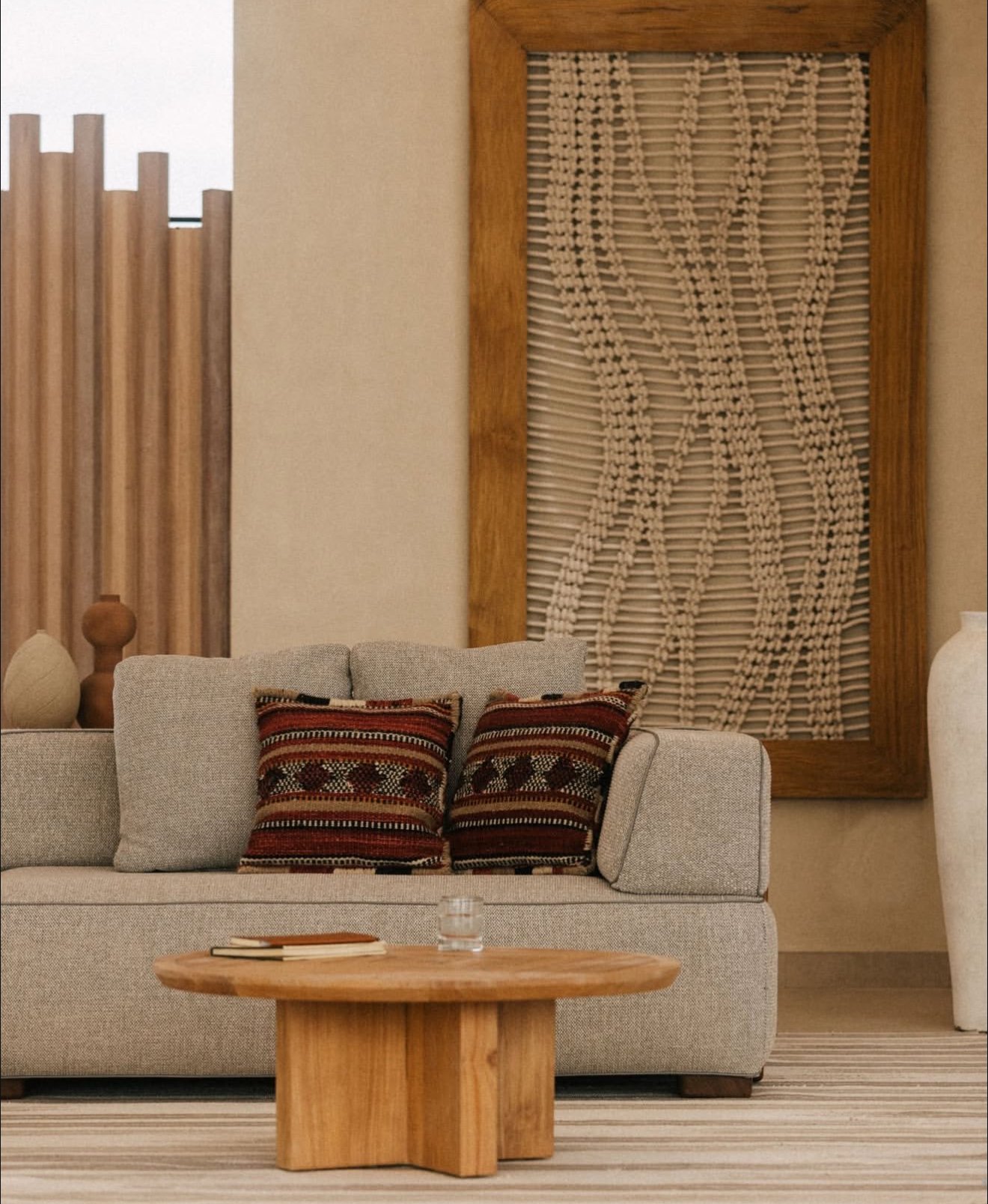 Living room with beige sofa, patterned throw pillows, wooden coffee table with books and glass of water, decorative wooden wall art, textured wall behind, striped rug.