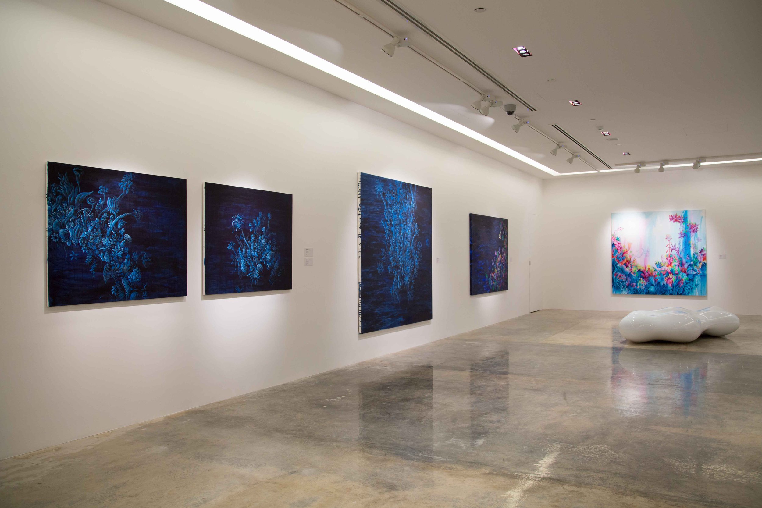 An art gallery with white walls displaying six vibrant paintings. Five paintings on the left feature predominantly blue floral designs, while the one on the right shows bright, colorful abstract floral art. A white, organic-shaped bench is positioned
