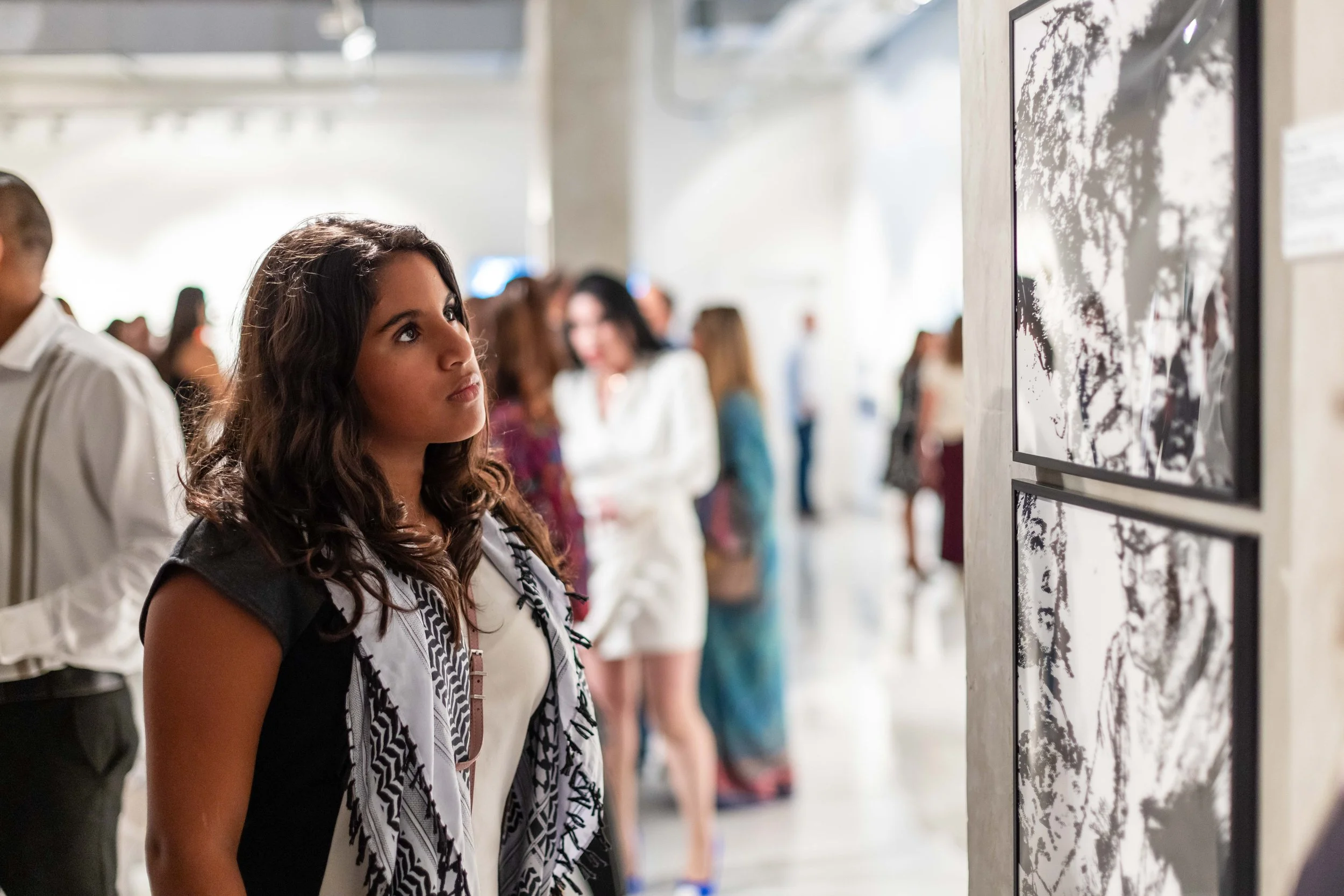 A young woman with long, curly dark hair looking at black and white abstract artwork in an art gallery with other visitors in the background.