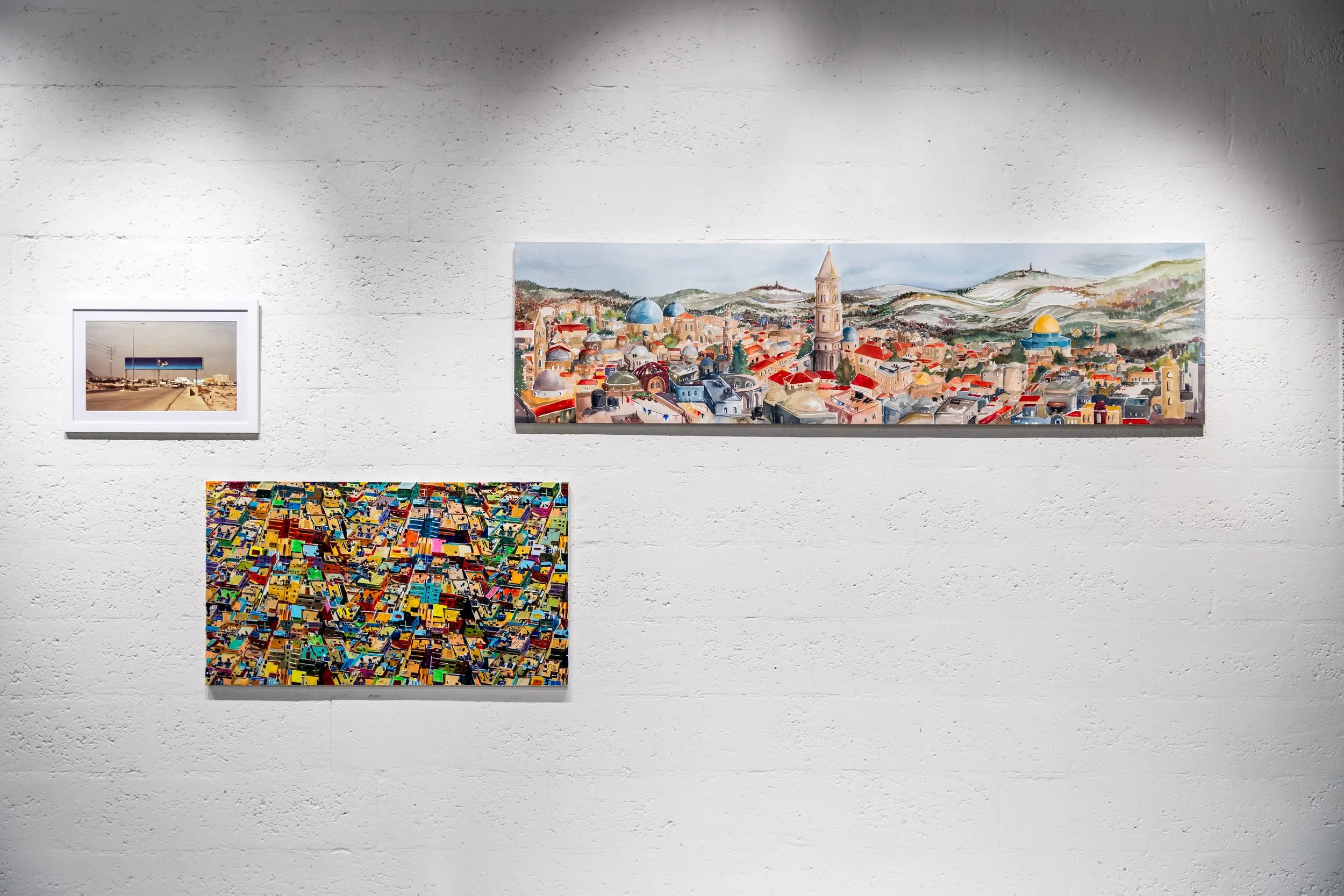 Three paintings on a white gallery wall: a small photograph of a desert landscape with power lines, a vibrant abstract of countless multicolored blocks, and a larger painting of a city skyline with buildings, domes, and hills in the background.