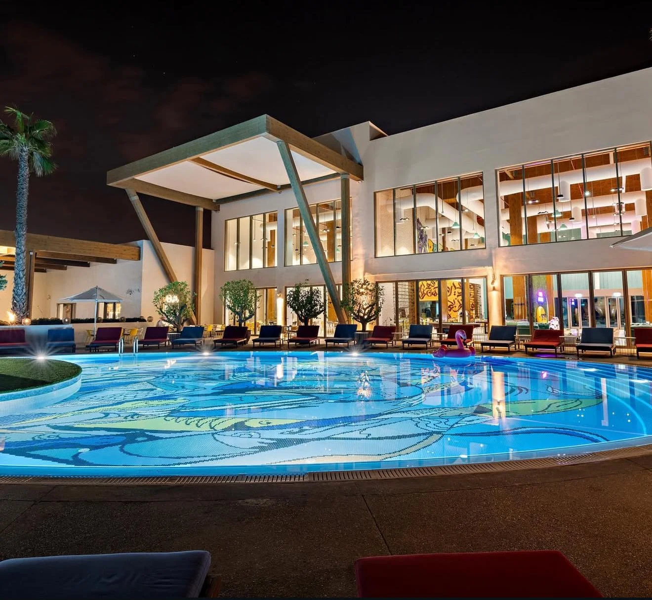 Night view of a luxury hotel pool with lounge chairs, an illuminated pool with a colorful design, and a modern building with large windows and interior lighting.