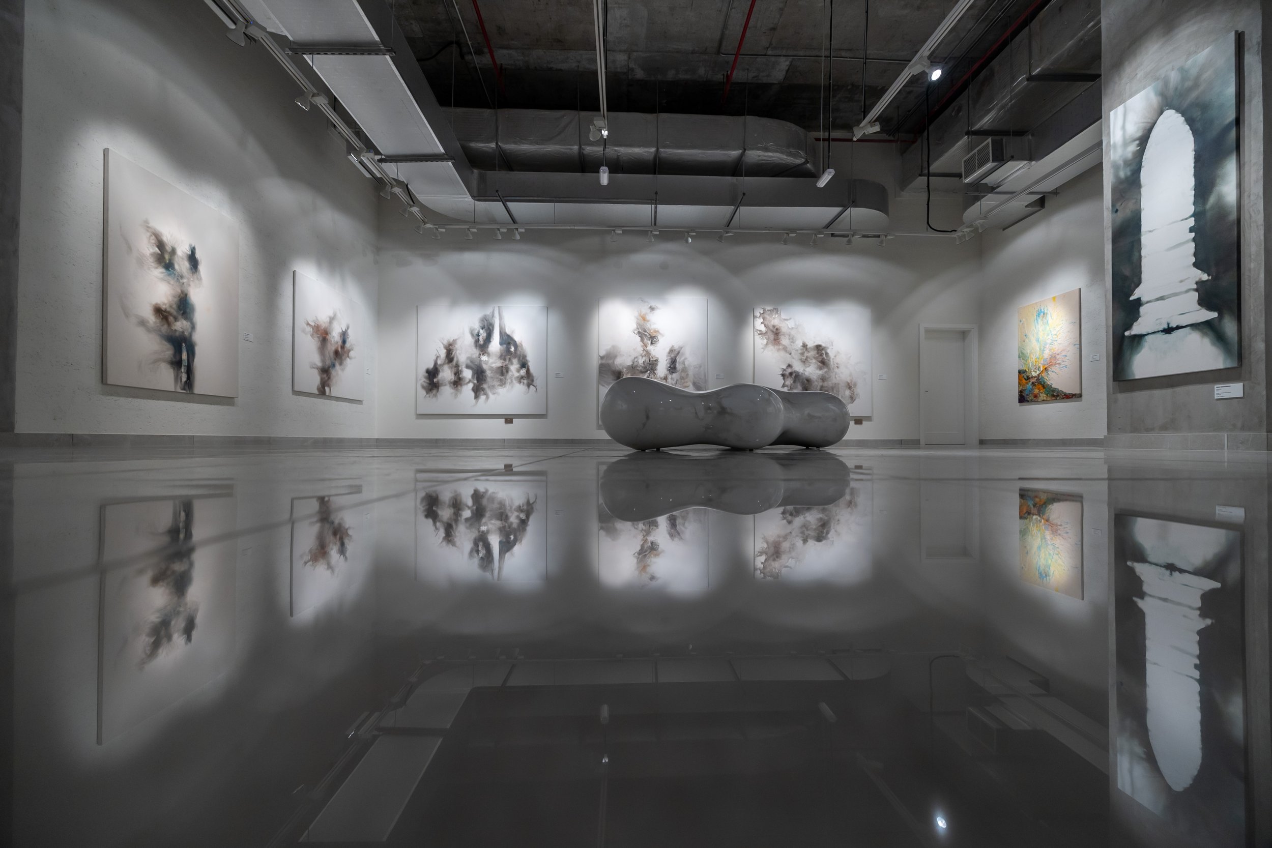 An art gallery featuring abstract paintings on white walls, a large reflective sculpture in the center, and a polished floor reflecting the artwork and sculpture.