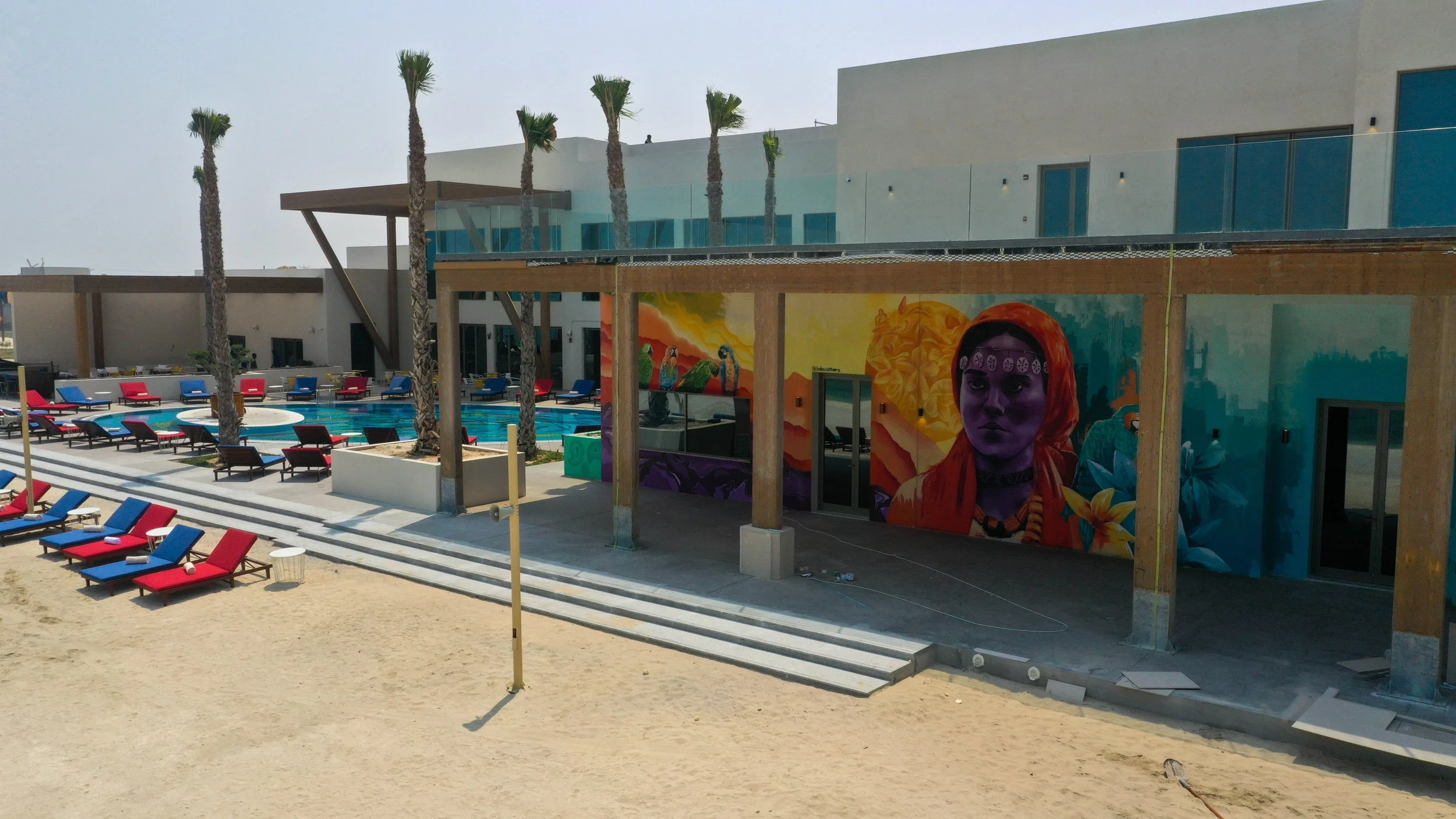 A modern beachside resort with a pool, colorful lounge chairs, and a vibrant mural on a building wall featuring a woman with an intense expression surrounded by floral and nature motifs.