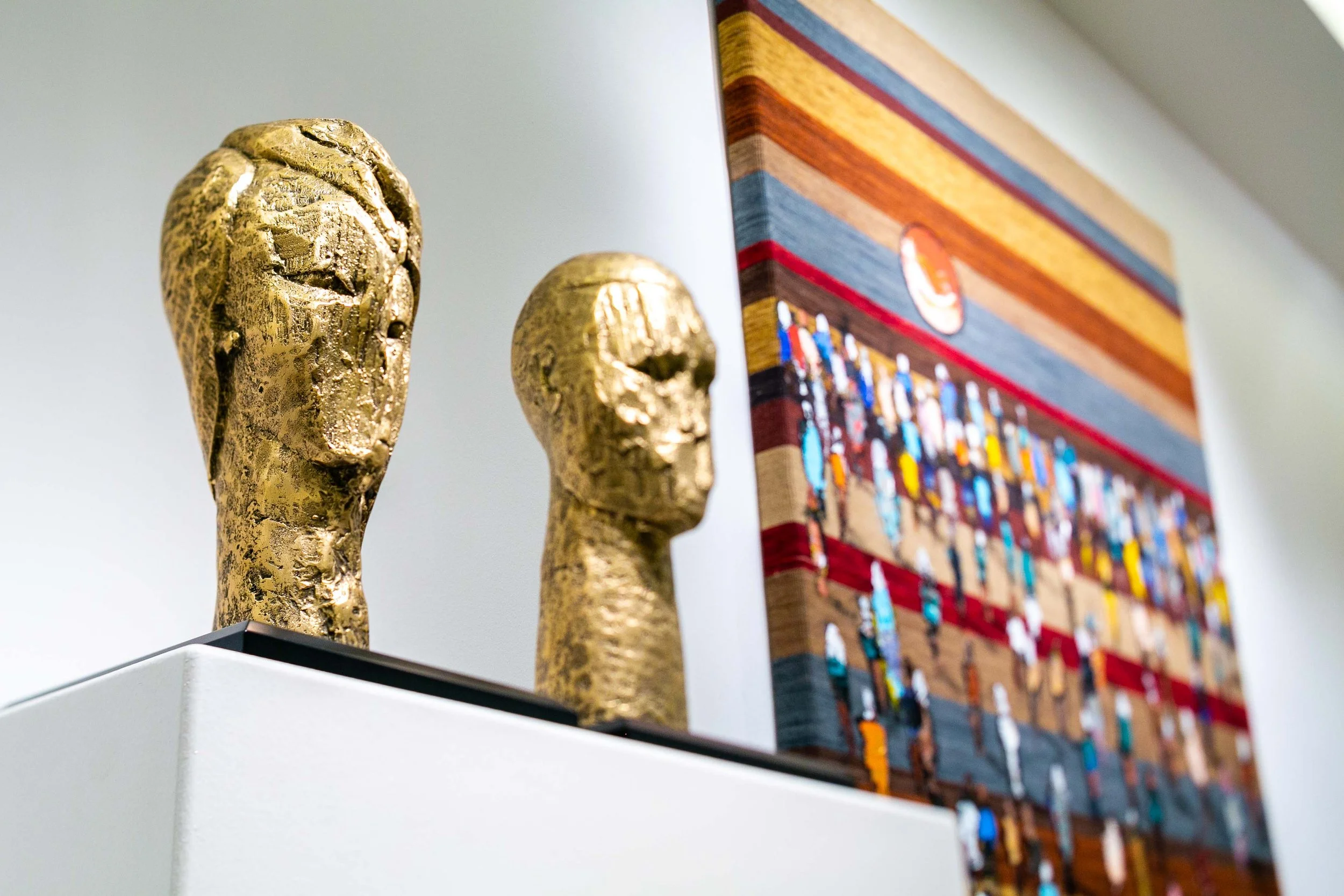 Close-up of two gold-colored sculptures of human heads on a white stand, with a colorful abstract artwork in the background.