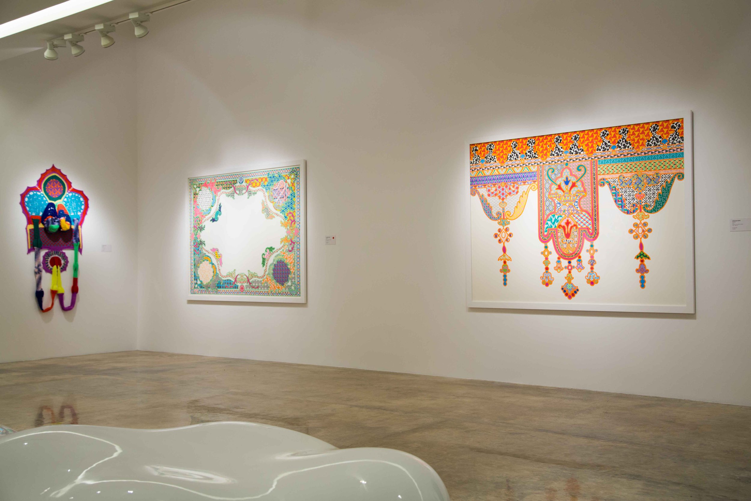 Contemporary art gallery with three brightly colored, patterned artworks displayed on white walls and a white sculpture in the foreground.