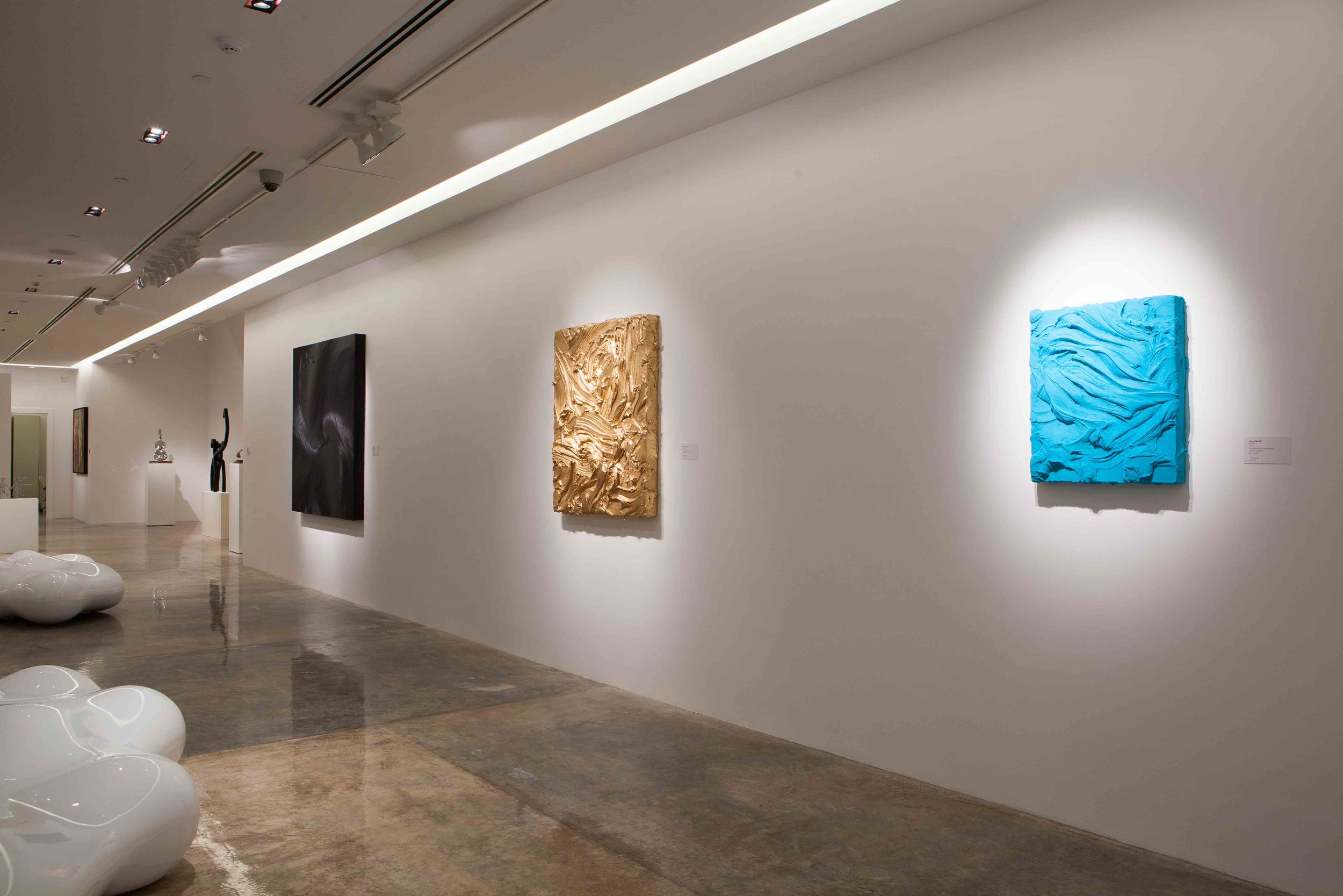 Inside an art gallery with white walls, featuring various abstract paintings and sculptures. There are glossy white oval seats on the floor in the foreground, and sculptures on pedestals along the left wall. Track lighting illuminates the artworks.