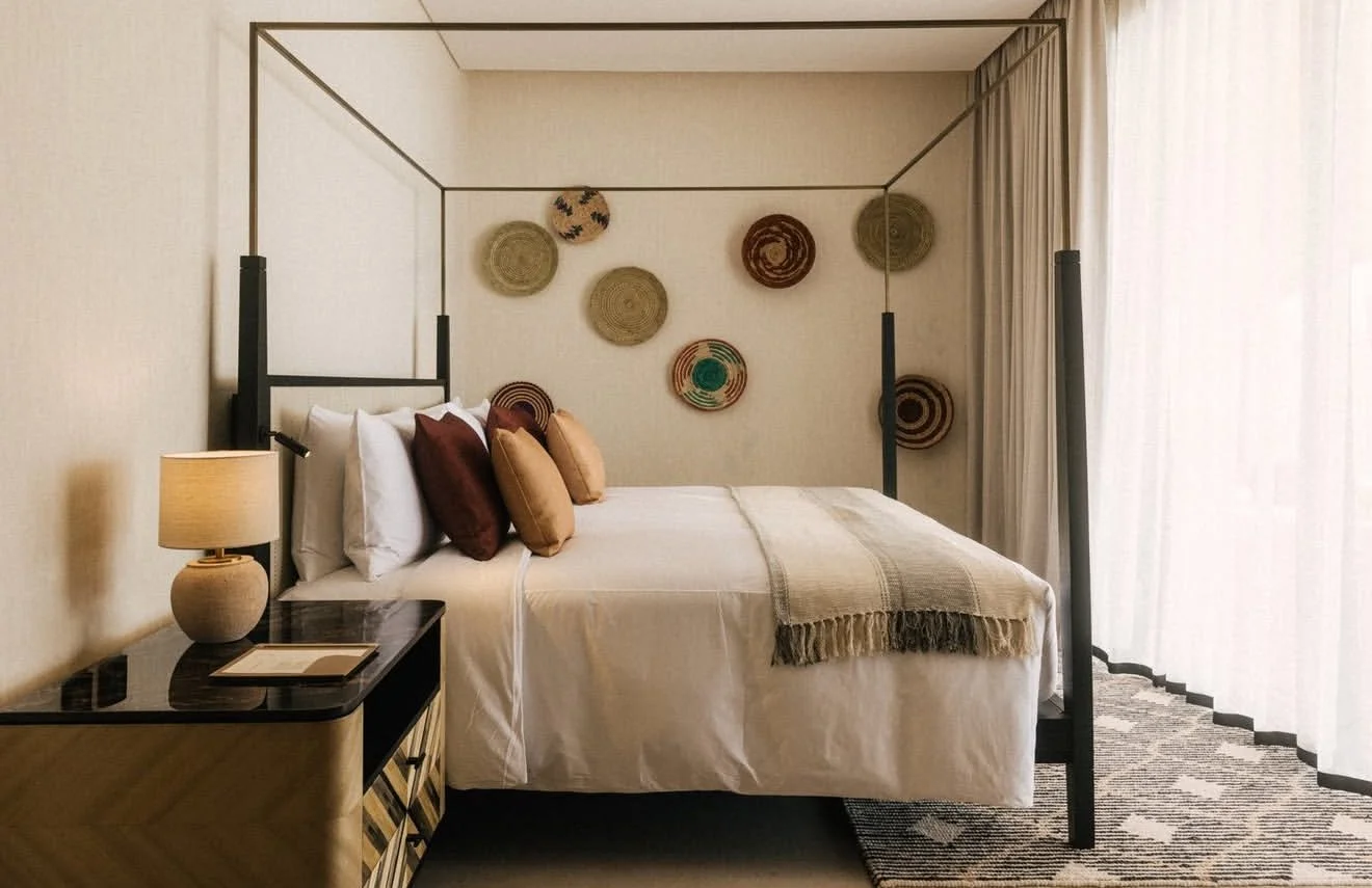 A bedroom with a four-poster bed, decorative pillows, a bedside table with a lamp, and woven wall hangings.