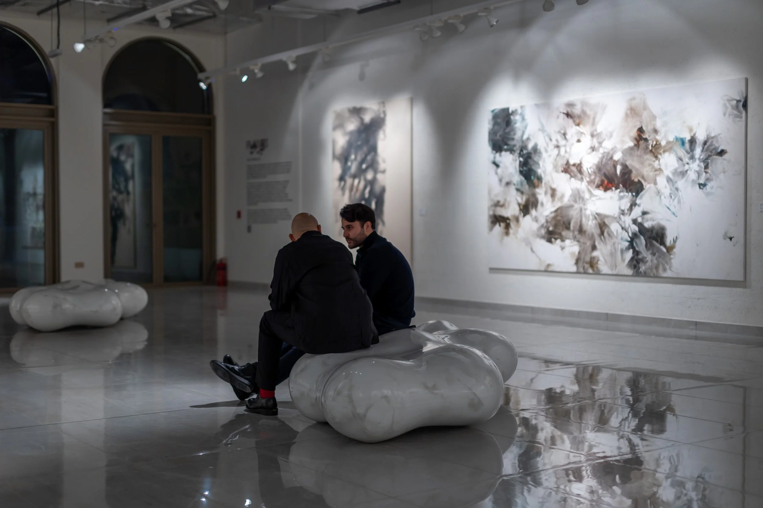 Two men sitting on a modern, glossy, white sculpture in an art gallery, with one facing away and the other looking at him. Abstract paintings hang on the wall behind them.